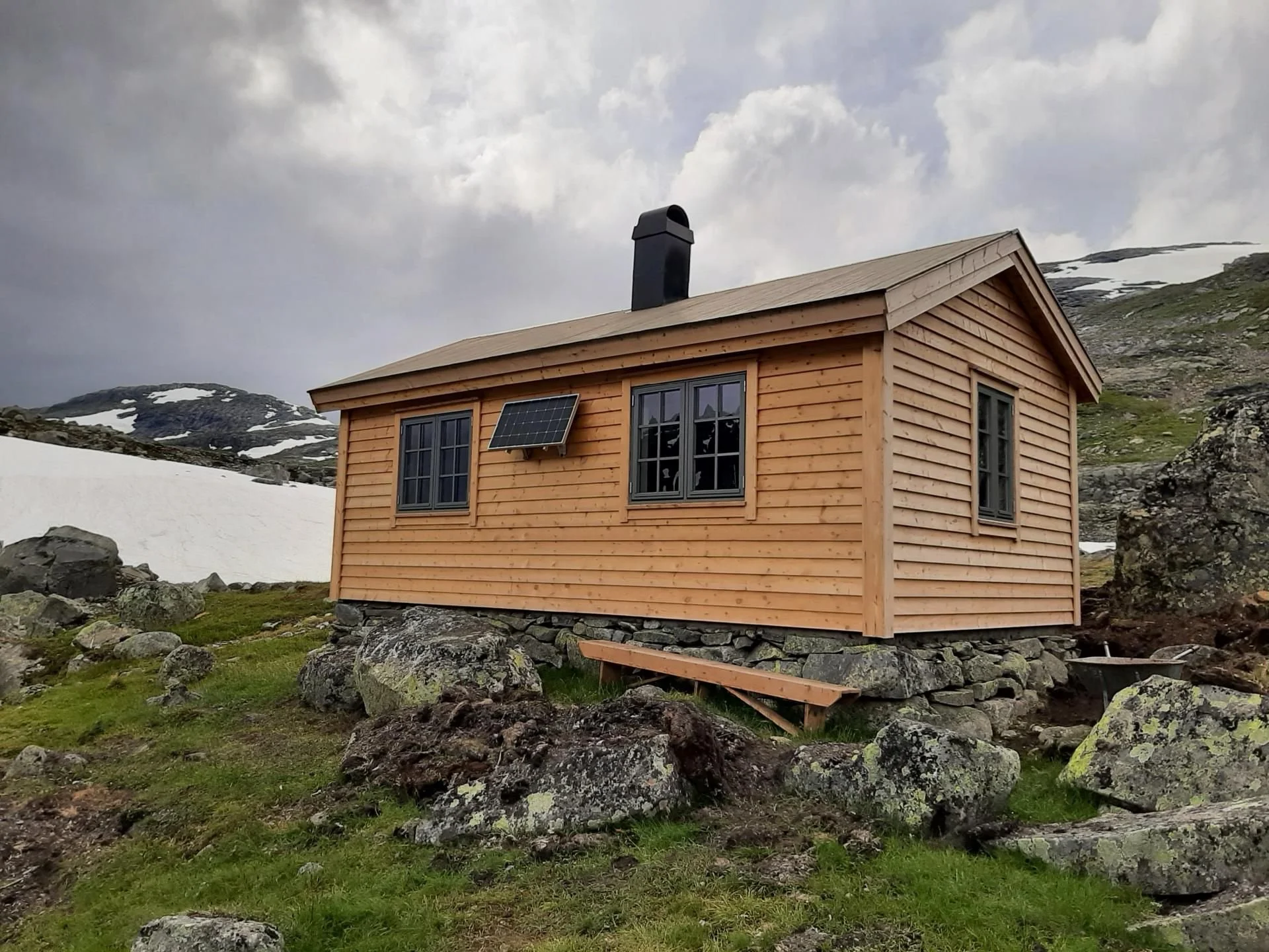 En liten trehytte med skifergrunnmur, omgitt av fjell med snø og steiner, under en overskyet himmel.