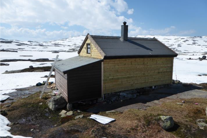 En liten tømret hytte med skråtak i et snødekt landskap.