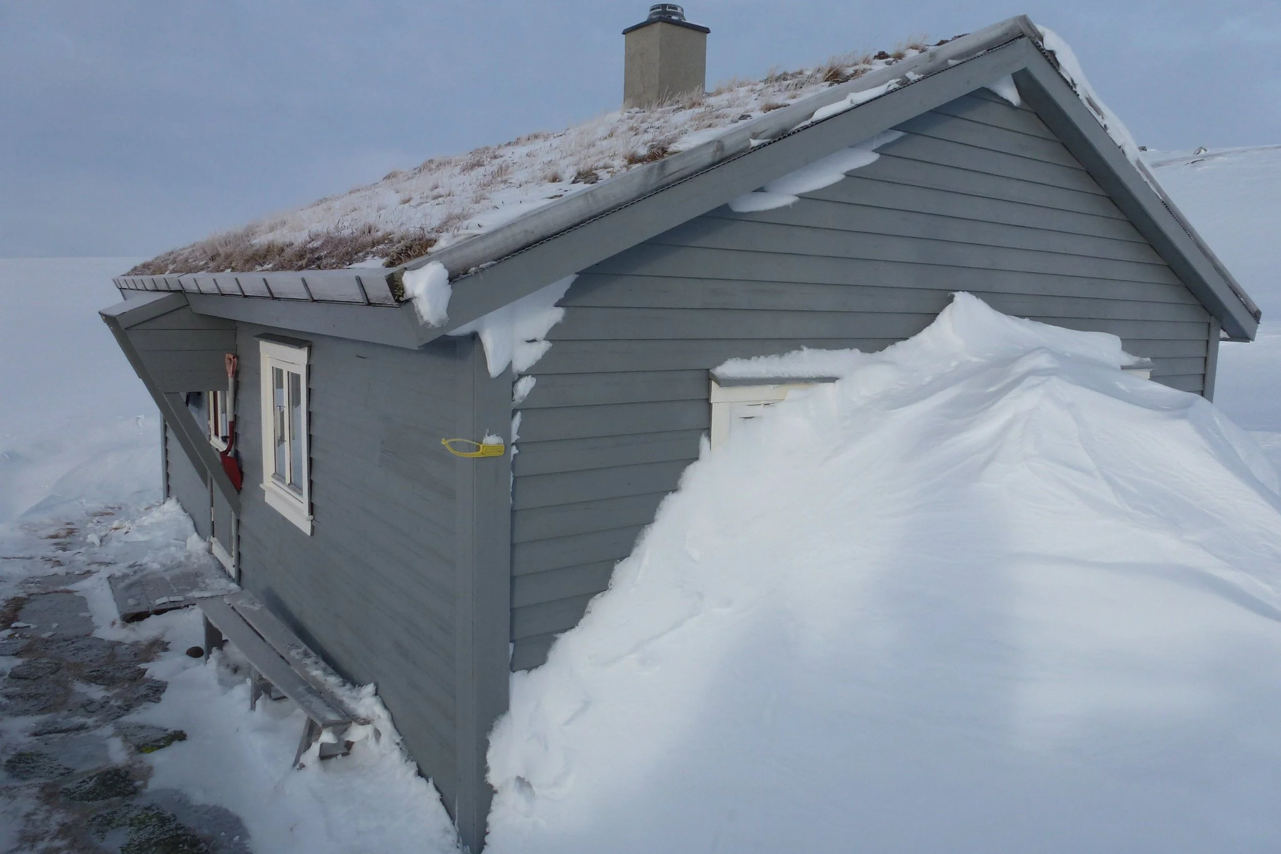 Et lite, grått hus omgitt av snø, med snøaya på taket og snø som har rast ned fra taket.