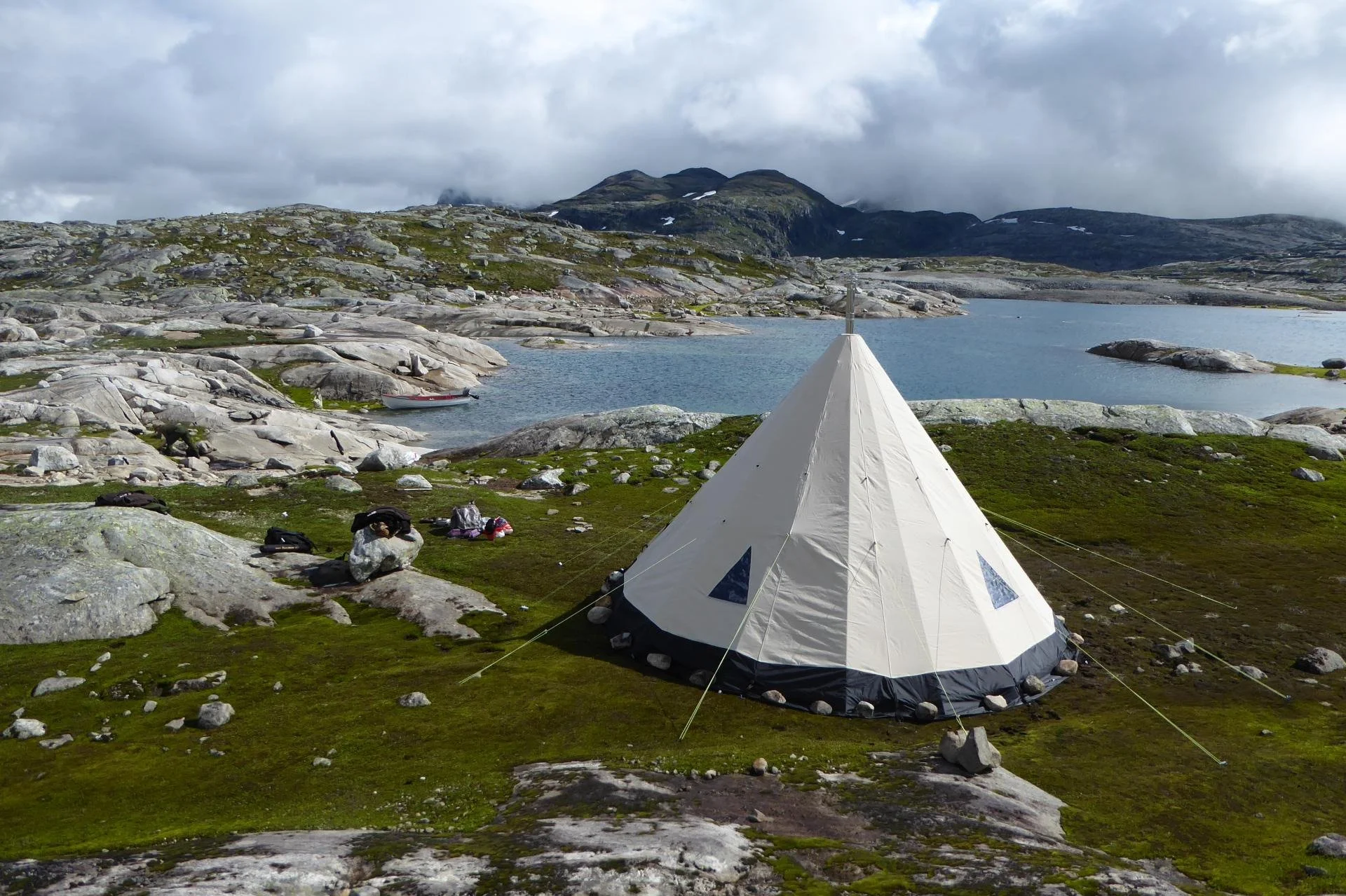 Telt satt opp ved en innsjø omgitt av store steiner og grønne myrer, med fjell i bakgrunnen under en overskyet himmel.