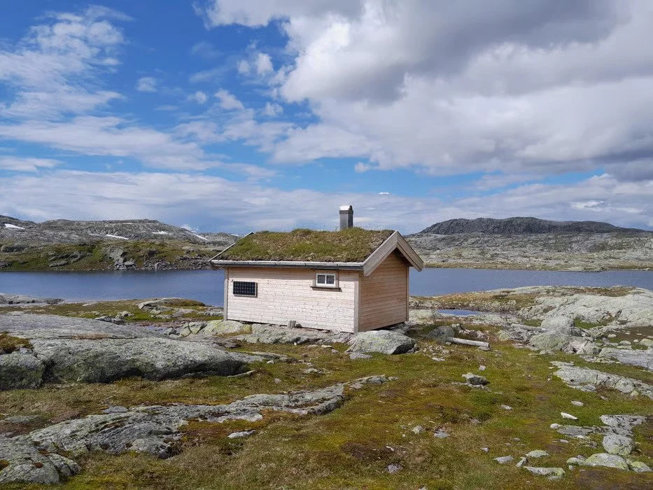 Liten hytte med grasdekt tak ved en innsjø i et fjellandskap under skyet himmel.