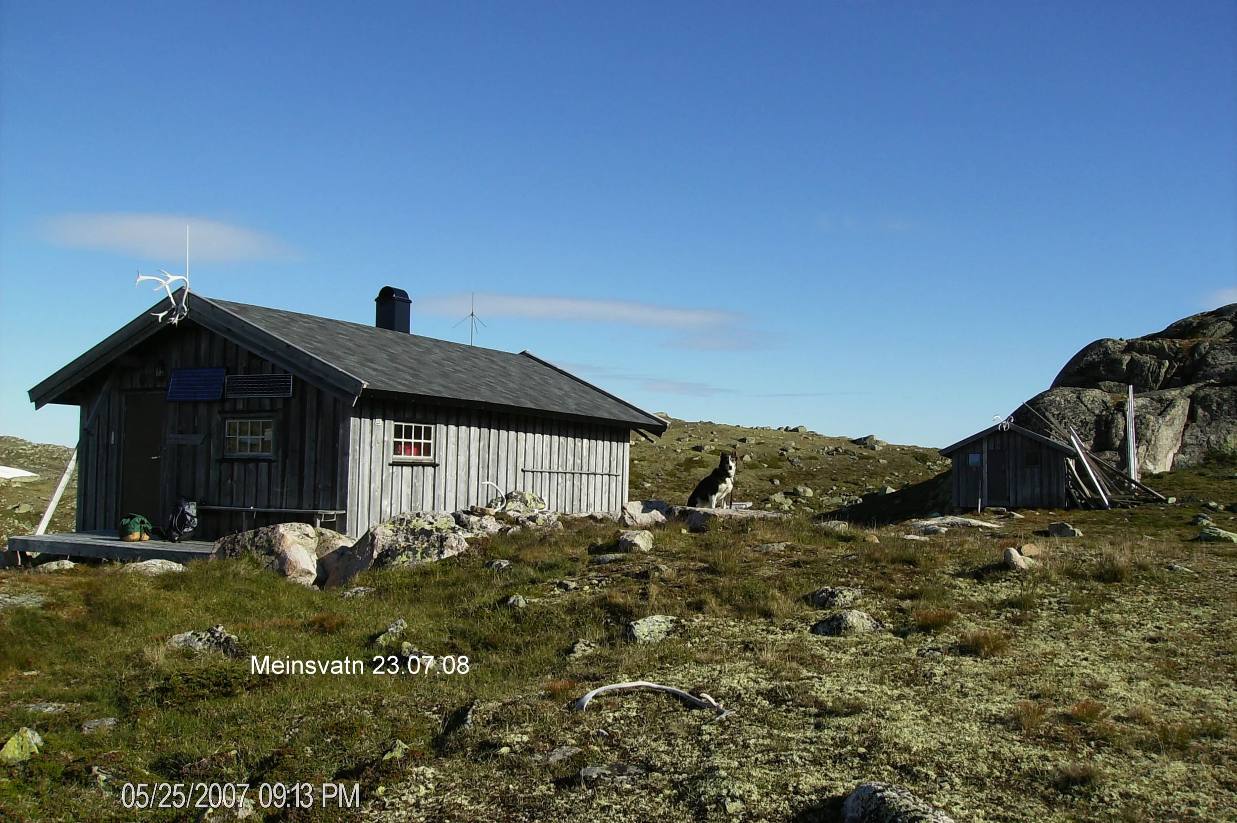 To små fjellhytter på en åpen, steinete slette med en hund som sitter mellom dem. Klar, blå himmel og noen skyer i bakgrunnen.