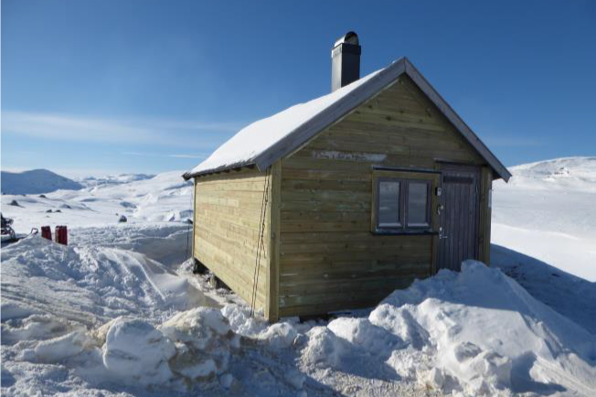 En liten trehytte dekket av snø i et snødekt landskap under en klar blå himmel.