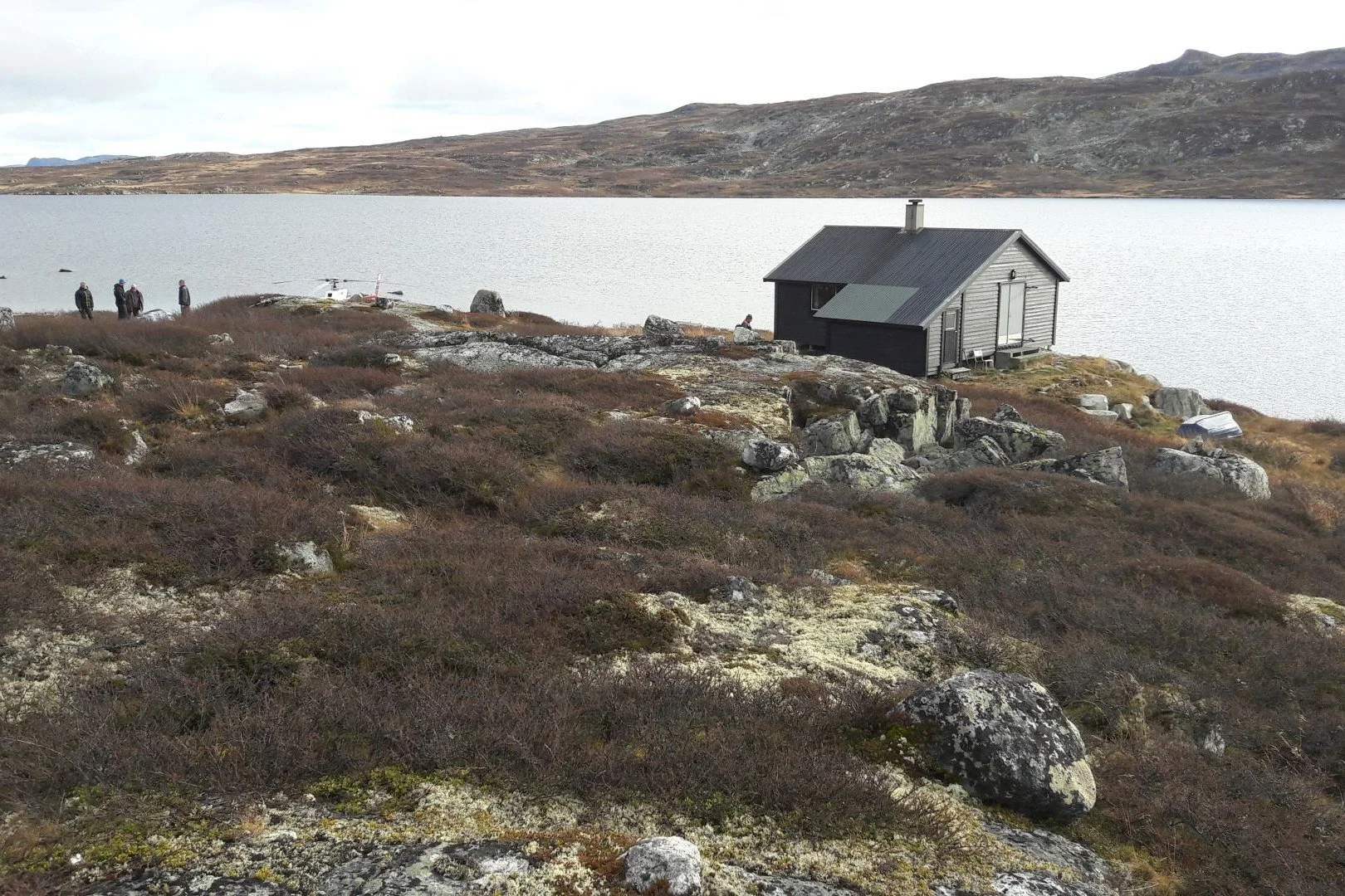 En liten svart hus med skråtak ved en innsjø omgitt av steinøse landskap og lav vegetasjon, med noen mennesker i bakgrunnen.