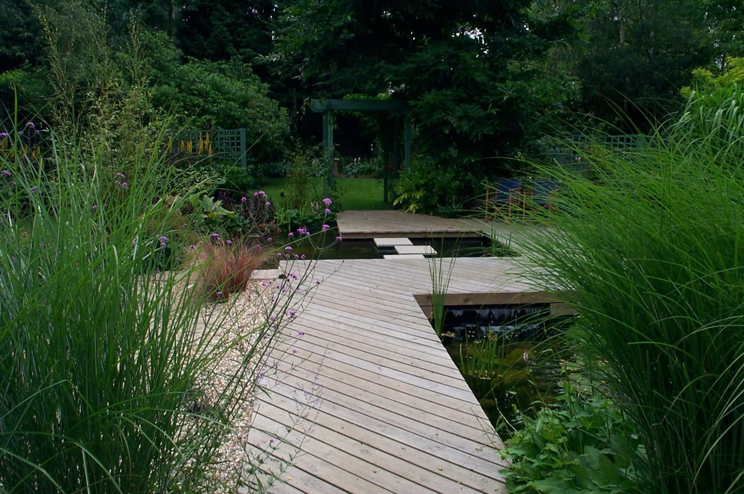 A wooden pathway leads through a lush green garden with tall grasses, purple flowers, and shrubs, towards a small pond and a dark green arbor in the background.