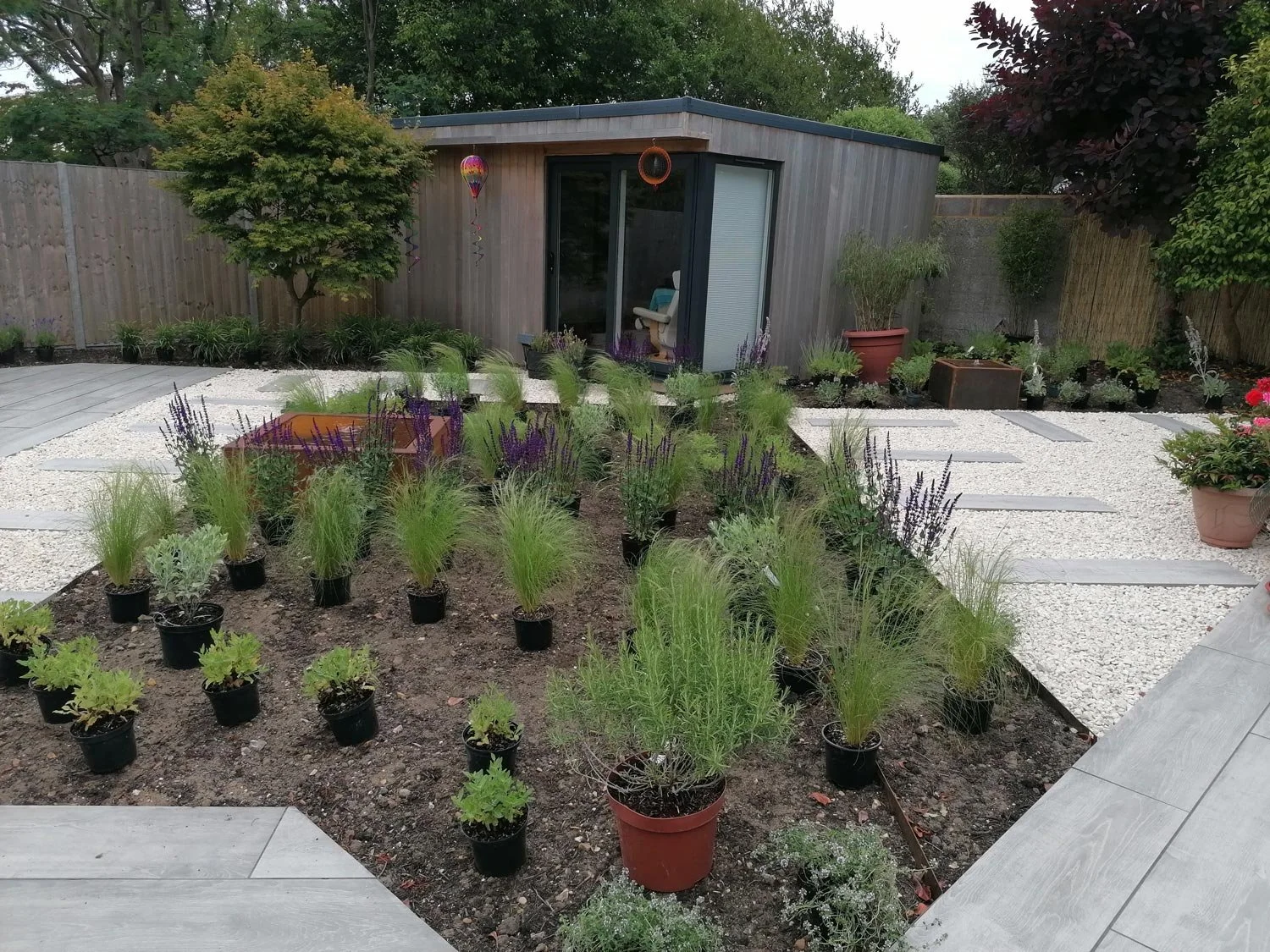 A backyard garden with potted plants, a small garden shed, a gravel pathway, and trees along the fence.