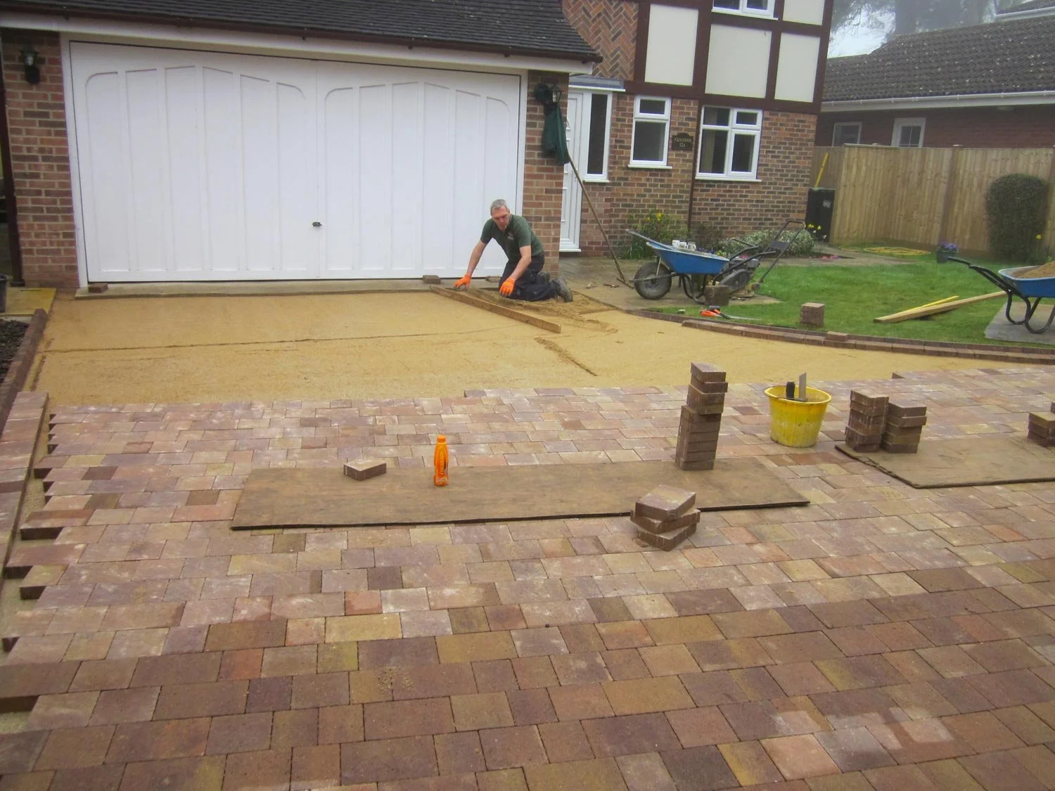 Person working on a landscaping project, laying new paving stones in a backyard, with wheelbarrows, building tools, and a yellow bucket nearby.