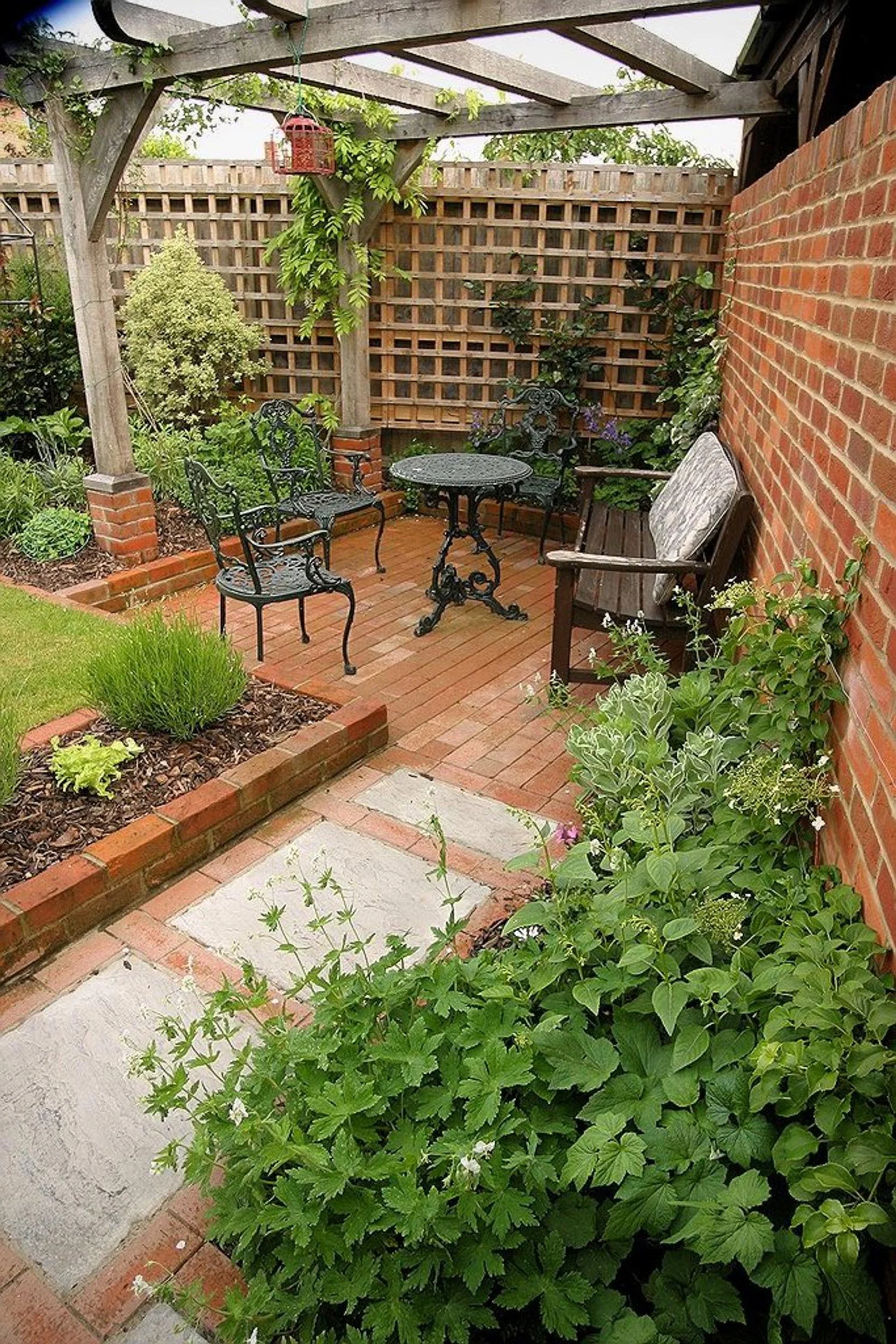 A small backyard garden patio with brick flooring, black wrought iron chairs and table, wooden bench, brick and wooden fencing, lush green plants, and a pergola with some greenery growing on it.