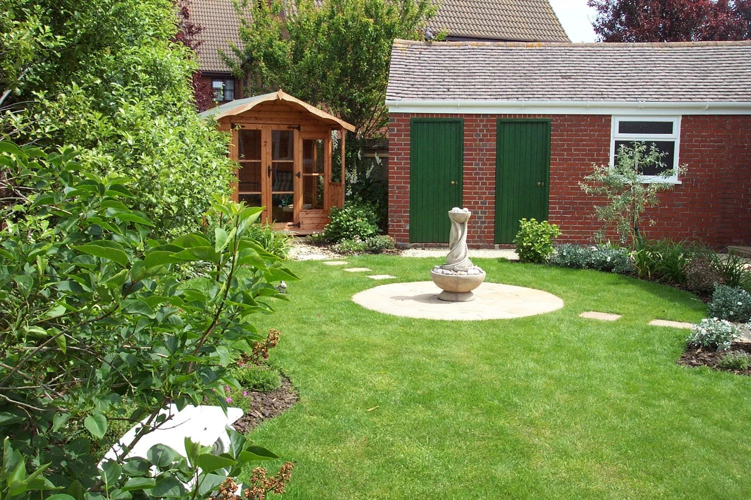 A well-maintained backyard garden with green grass, a birdbath in the center, and lush bushes on the sides. In the background, there is a small wooden garden shed with a curved roof and glass doors, next to a brick building with two green doors and a