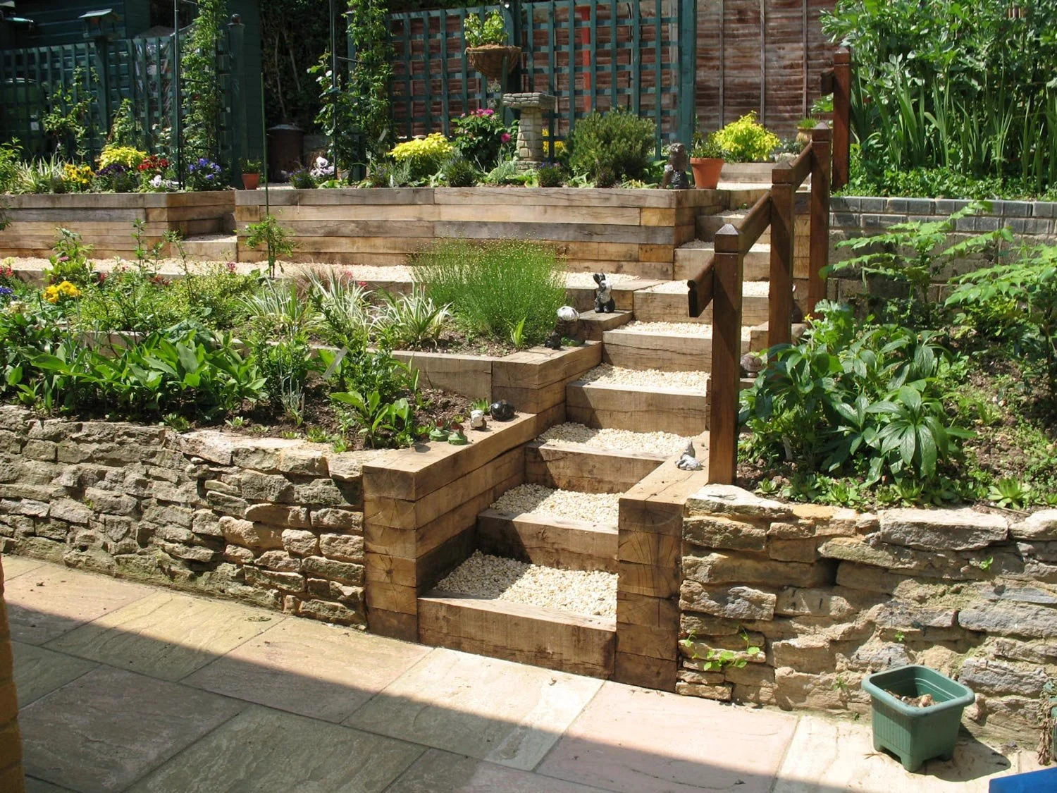 A tiered garden with stone and wooden steps, lush green plants, and flowers, surrounded by a stone wall and fencing.