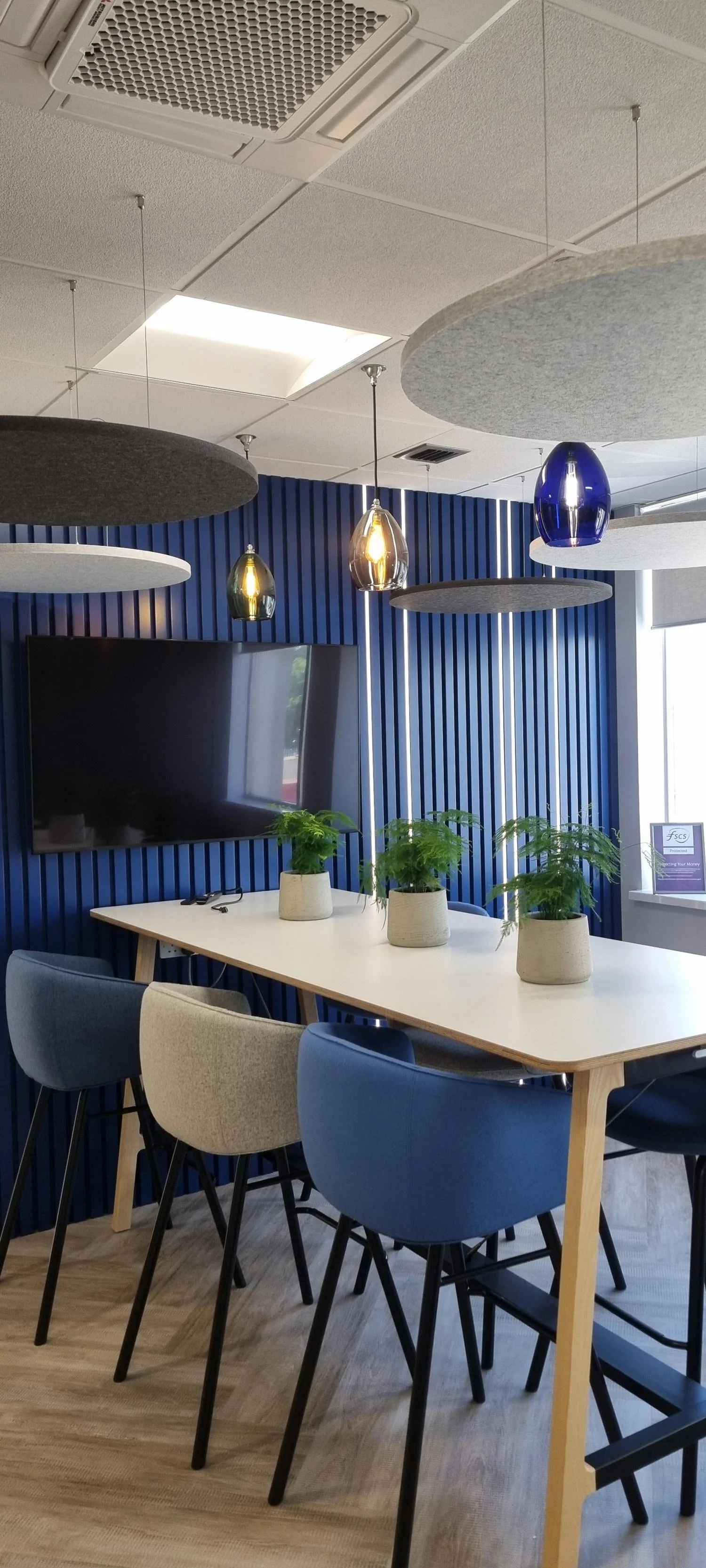 Modern interior space with a white table decorated with three potted plants and six chairs around it, against a blue textured wall with a large screen, a window, hanging lights, and ceiling panels.