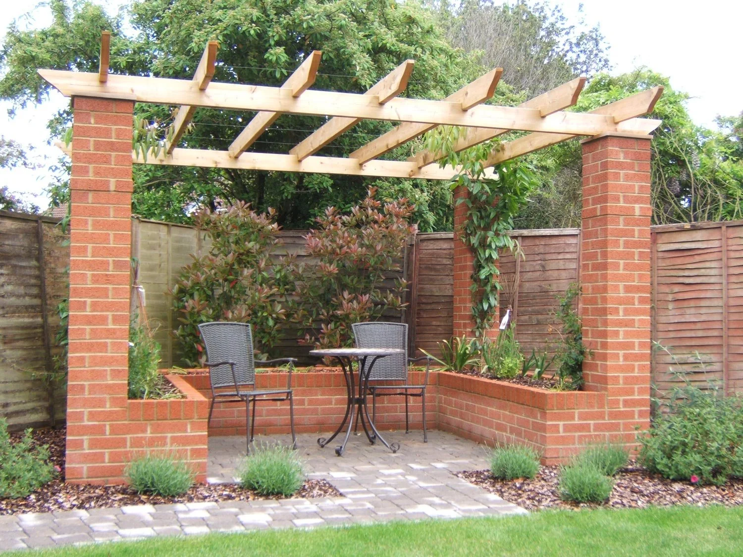 Backyard patio with brick garden wall, firepit table, two chairs, and a wooden pergola frame.