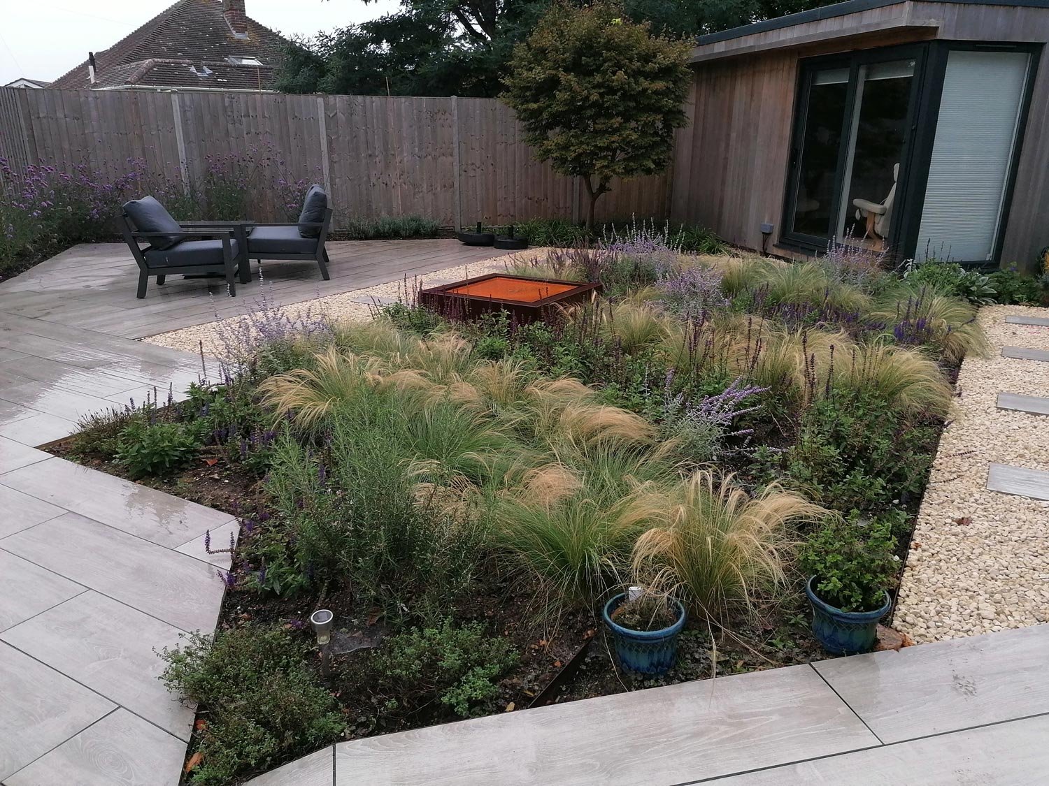 A modern backyard garden with a circular bed of ornamental grasses and purple flowering plants, bordered by light-colored paving stones and a gravel pathway. There is a small fire pit in the center of the garden, and a seating area with two black arm