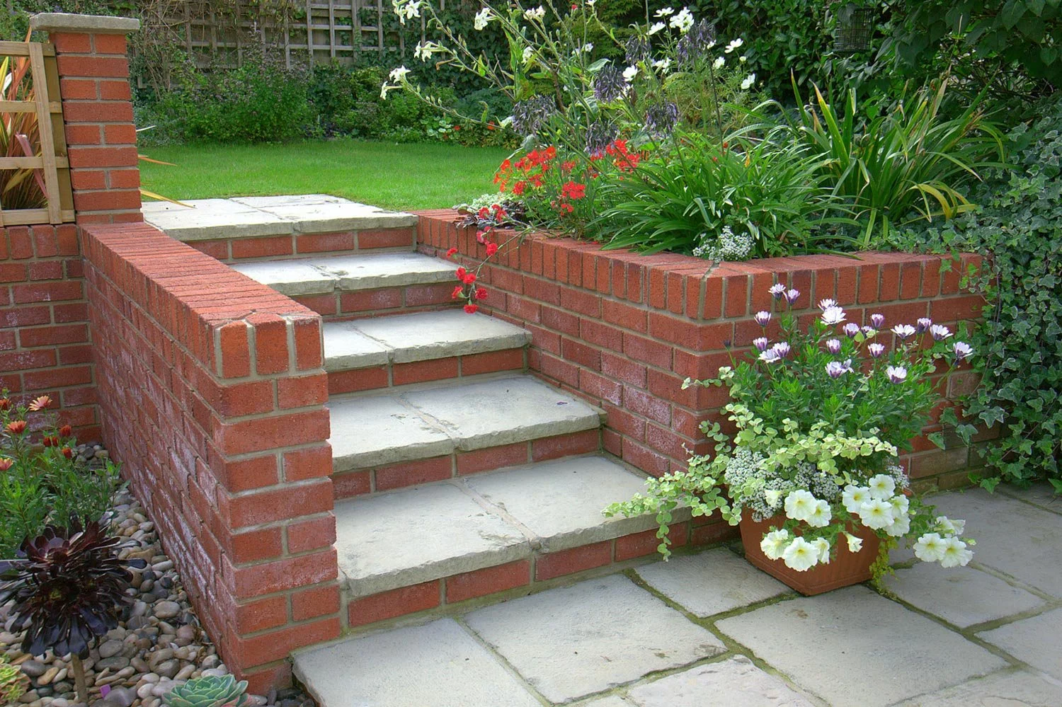 A garden with brick steps leading to a grassy lawn, surrounded by a brick wall with lush green plants and colorful flowers in a planter.