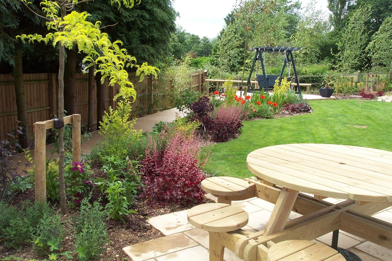 A backyard garden featuring a wooden picnic table, a well-manicured green lawn, flowering plants, a wooden fence, and a swing set in the distance.