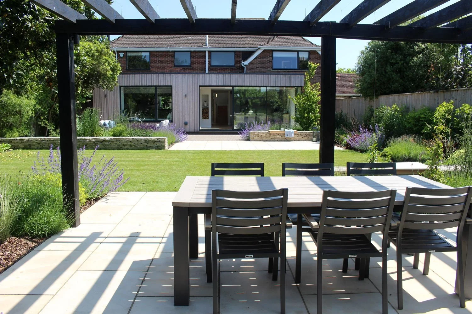 A backyard patio with a dining table and chairs under a pergola, overlooking a well-maintained lawn and garden with various plants and shrubs, and a modern two-story house in the background.