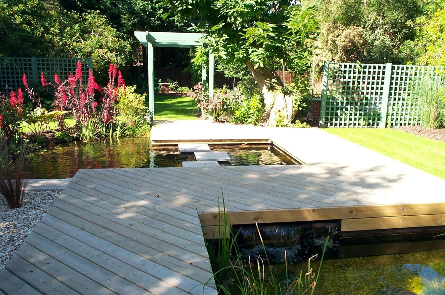 A backyard garden featuring a wooden deck, a pond with floating stepping stones, surrounded by lush green plants and colorful flowers, with a pergola and trees in the background.