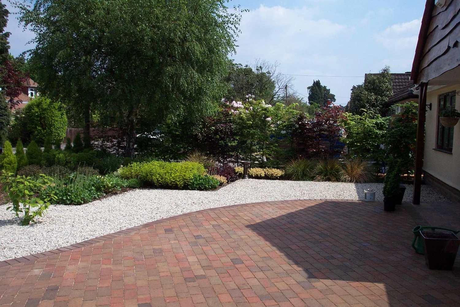 A backyard with a brick patio, a garden with various green plants, bushes, and trees, and a gravel pathway separating the patio from the garden. There are some potted plants and gardening tools visible.
