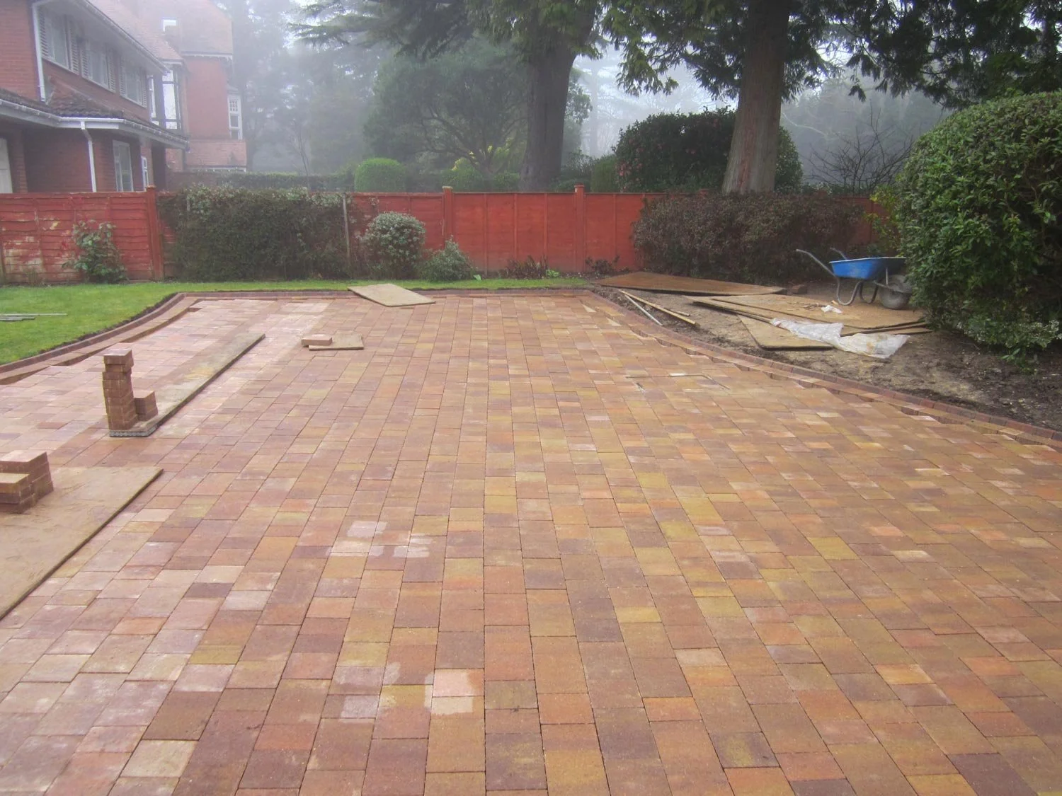 A backyard with a newly paved brick patio, construction materials, and tools, with a red fence, bushes, and trees in the background.