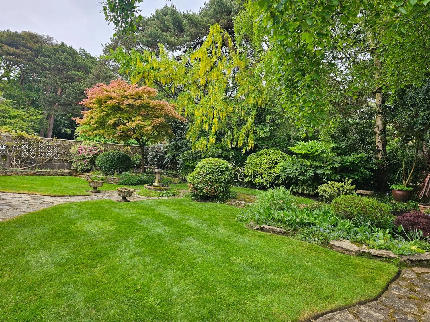 A lush garden with a well-manicured green lawn, surrounded by various trees, shrubs, and flowering plants, with a stone pathway and decorative pots.