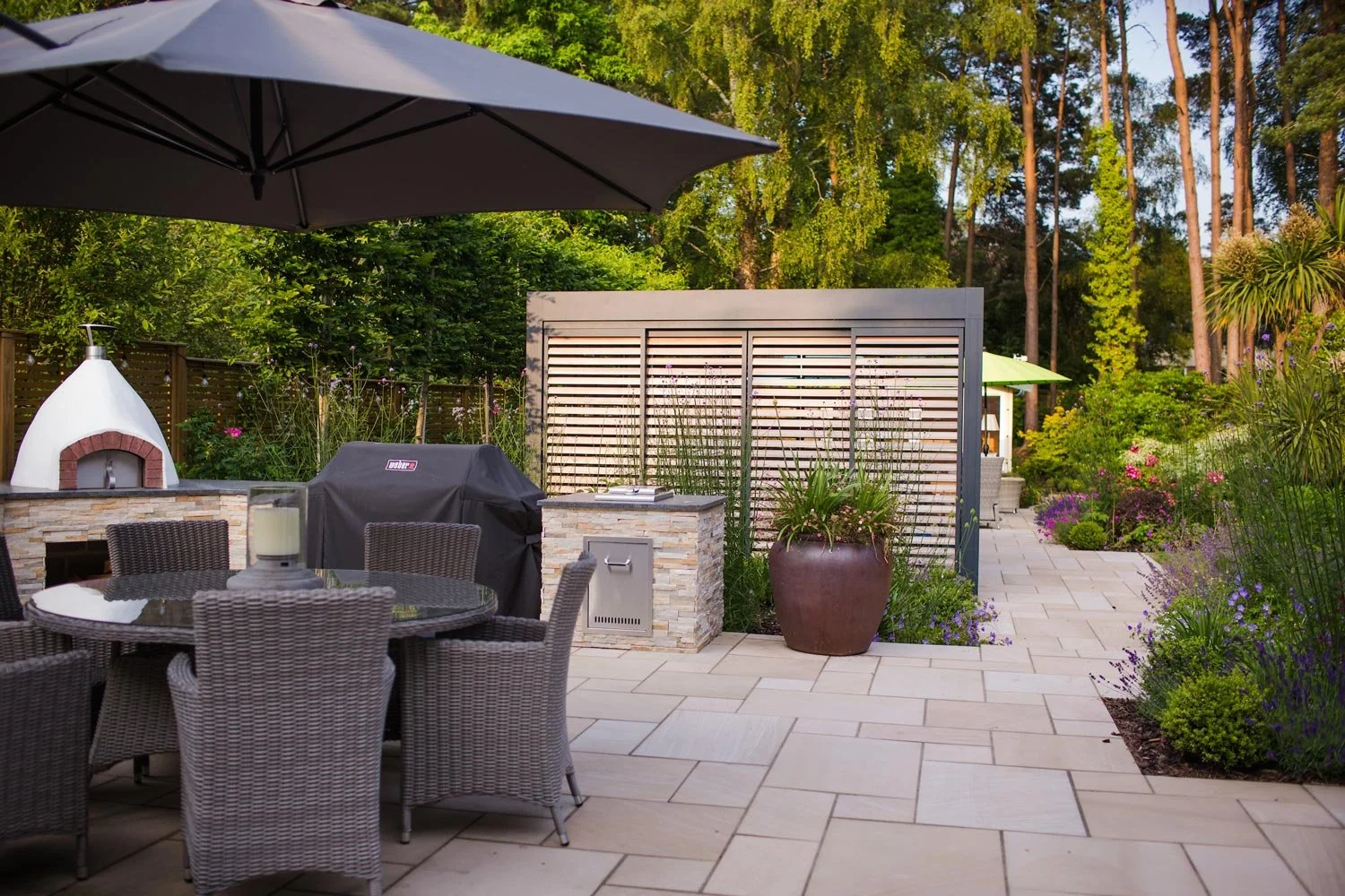 Outdoor patio with a black umbrella, dining table with chairs, a pizza oven, covered grill, potted plants, and a landscaped garden with flowers and trees.