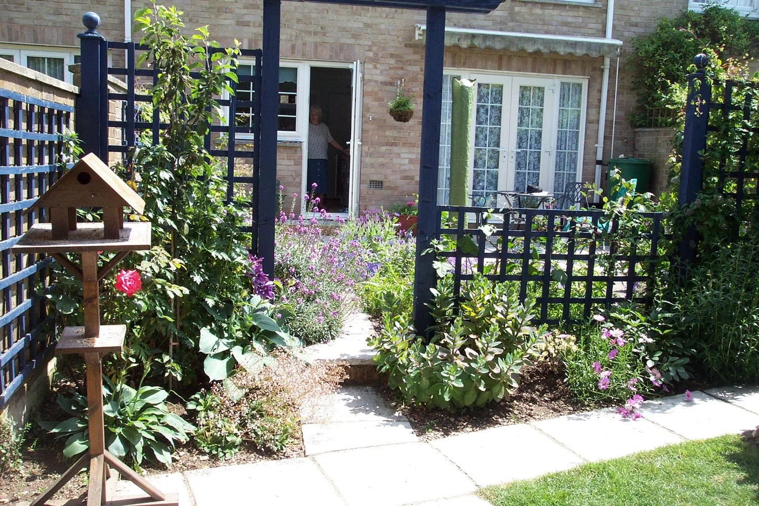 A backyard garden with flowering plants, a wooden birdhouse on a stand, a paved pathway, and a woman standing in the doorway of a brick house, visible through an open door.