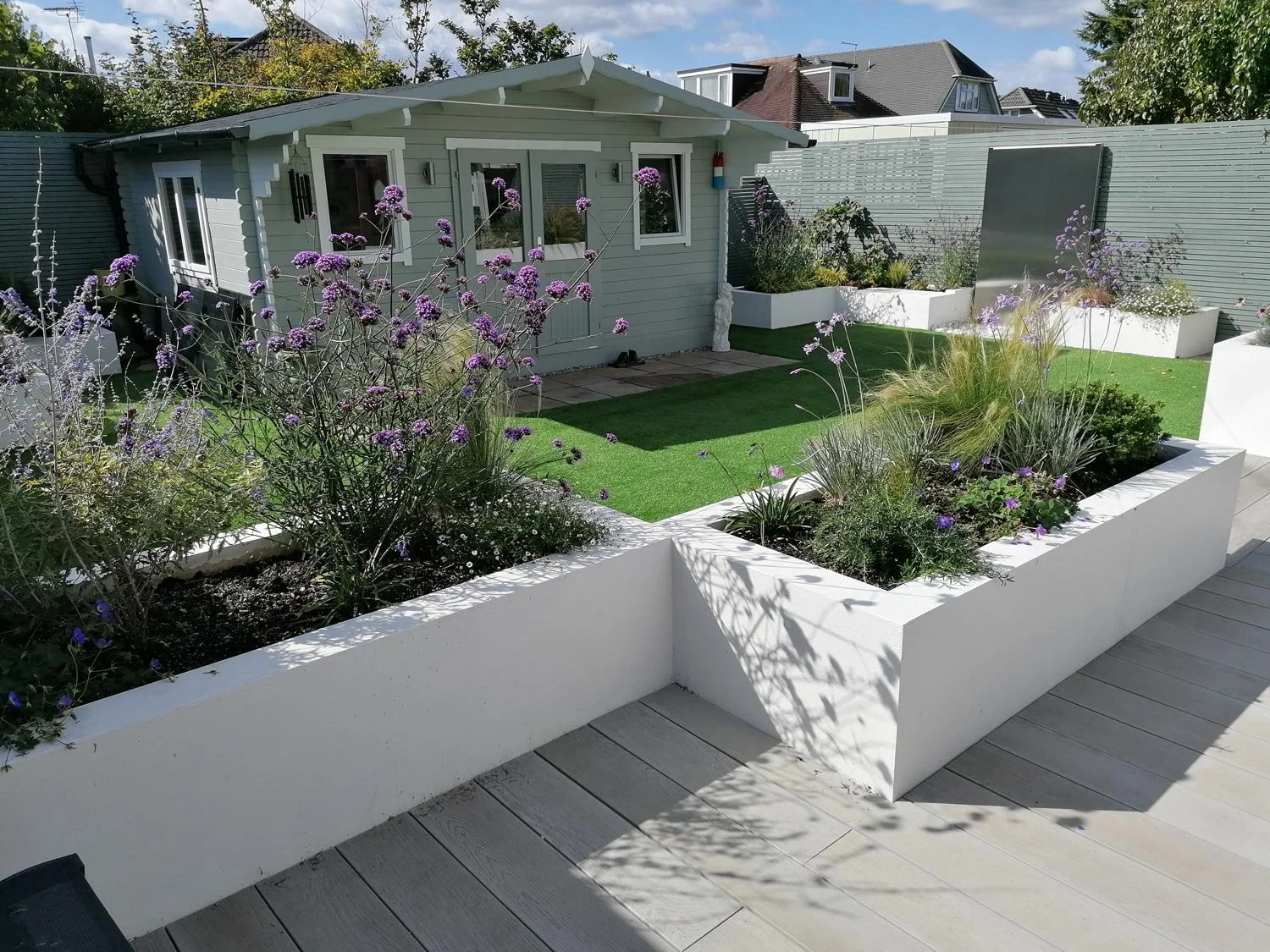 A backyard garden with raised white flower beds filled with purple flowers and grasses, a small patch of artificial green turf, and a small house painted in light blue with white trim.