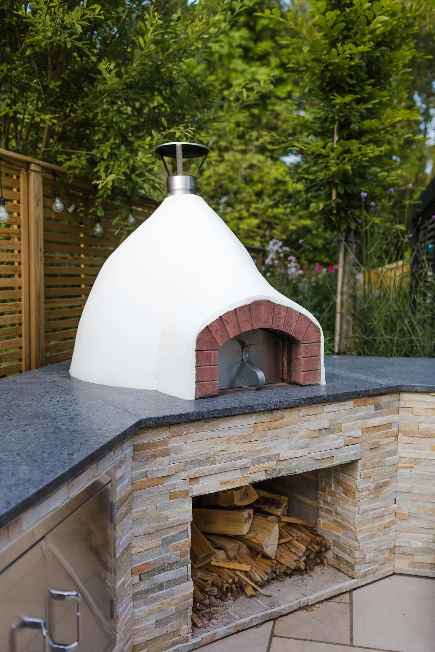 Outdoor pizza oven with a white dome and brick arch, rest on a stone counter with a wood storage space below, surrounded by greenery and string lights.