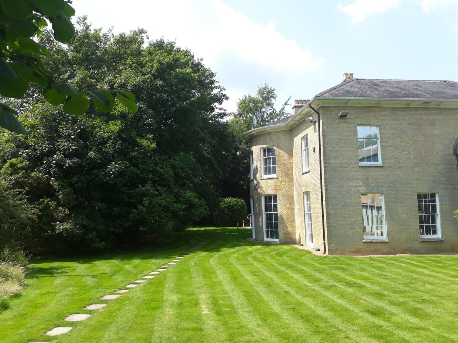 Large brick house with multiple windows and a curved exterior wall, surrounded by a well-maintained, striped green lawn and tall trees.