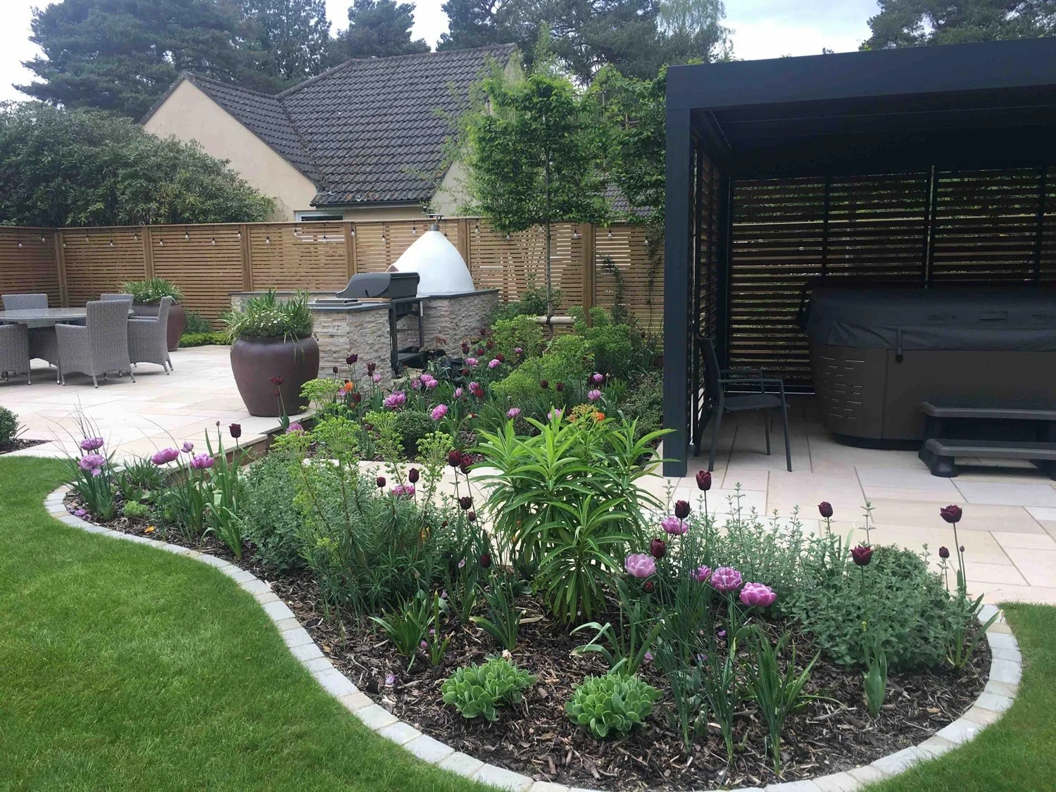 A backyard garden with a curved flower bed filled with purple and dark red flowers, surrounded by a neatly manicured lawn, and features outdoor furniture, a barbecue grill, a hot tub under a protective cover, and a privacy fence, with trees and a house in the background.