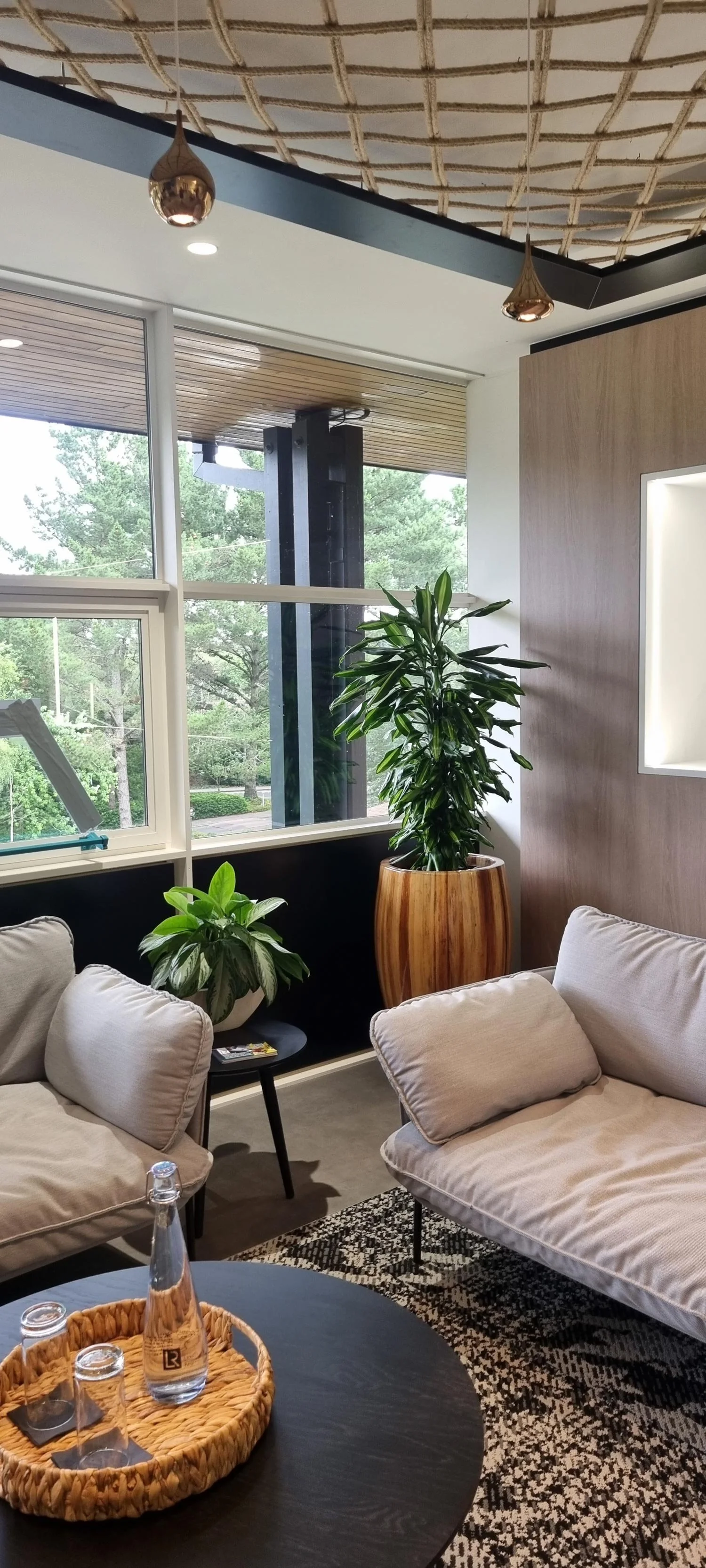 A cozy living room with beige sofas, green potted plants, a black round table with a wicker tray holding a glass water bottle and glasses, large windows with greenery outside, and decorative metallic pendant lights hanging from a patterned ceiling.