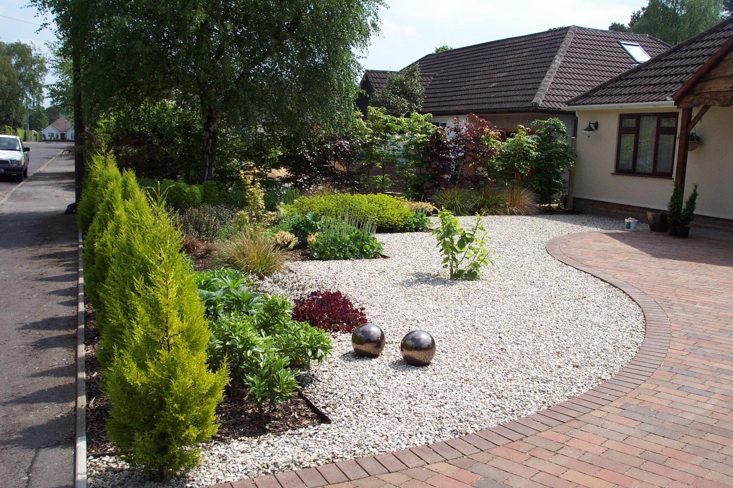 A landscaped front yard with small shrubs, plants, and a gravel area surrounded by a curved brick walkway. There are two metallic spheres on the gravel, with houses and trees in the background.