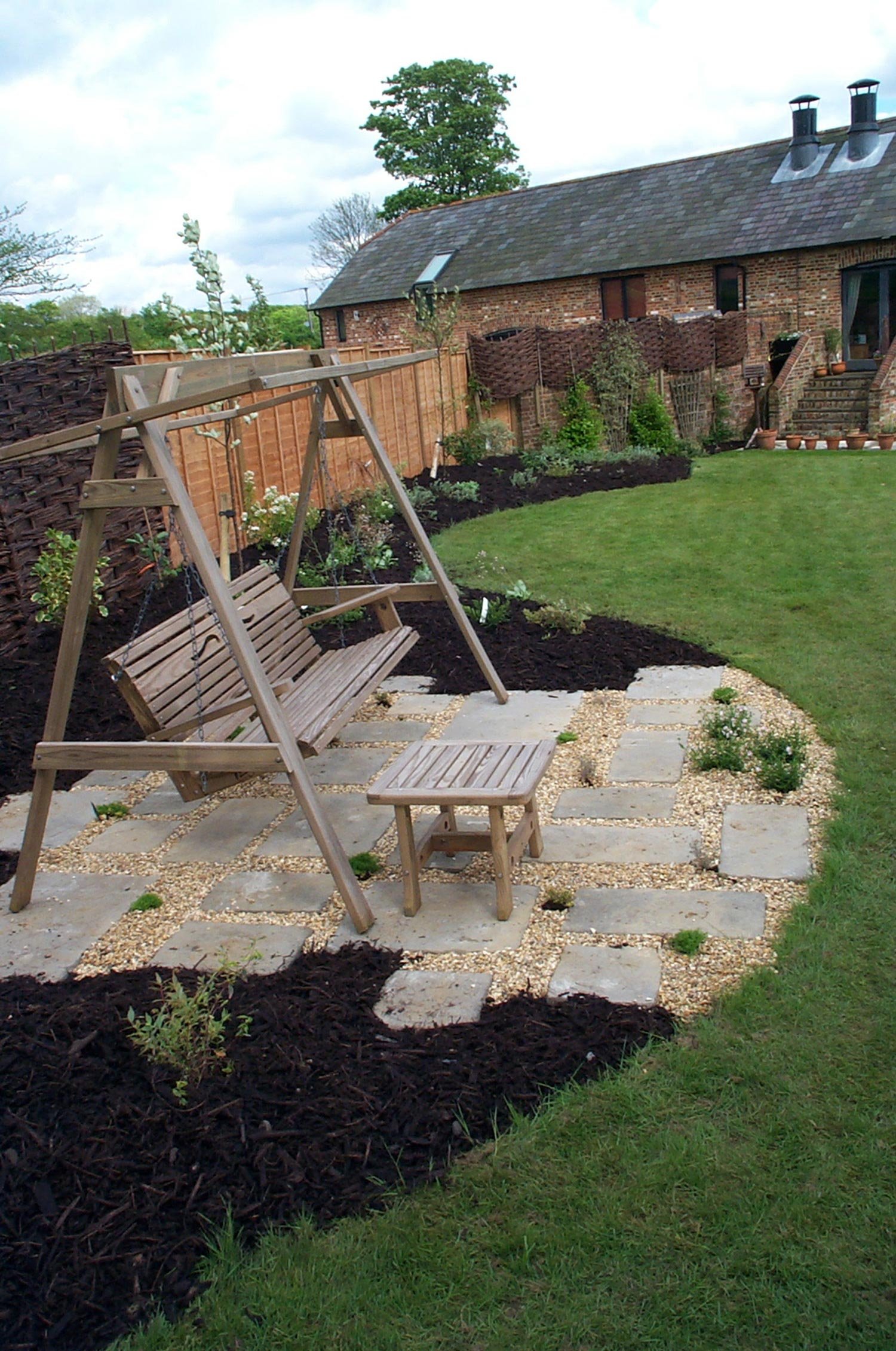 A backyard garden with a wooden swing bench on a stone and gravel patio, adjacent to green grass and landscaped flower beds with plants and bushes, enclosed by a wooden fence and open sky.
