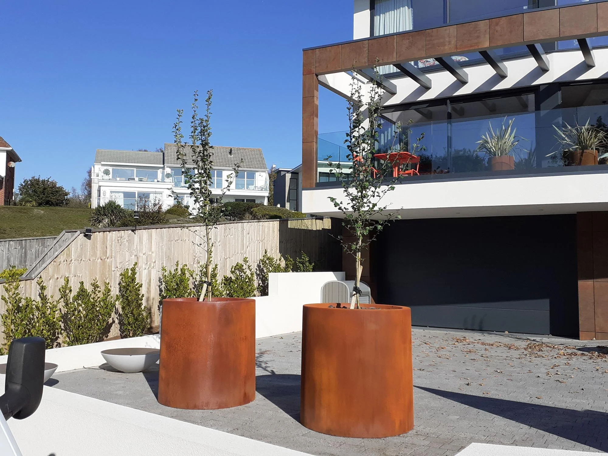 Modern residential building with glass balcony, outdoor plants, and two rust-colored planters with small trees in a paved courtyard under a clear blue sky.