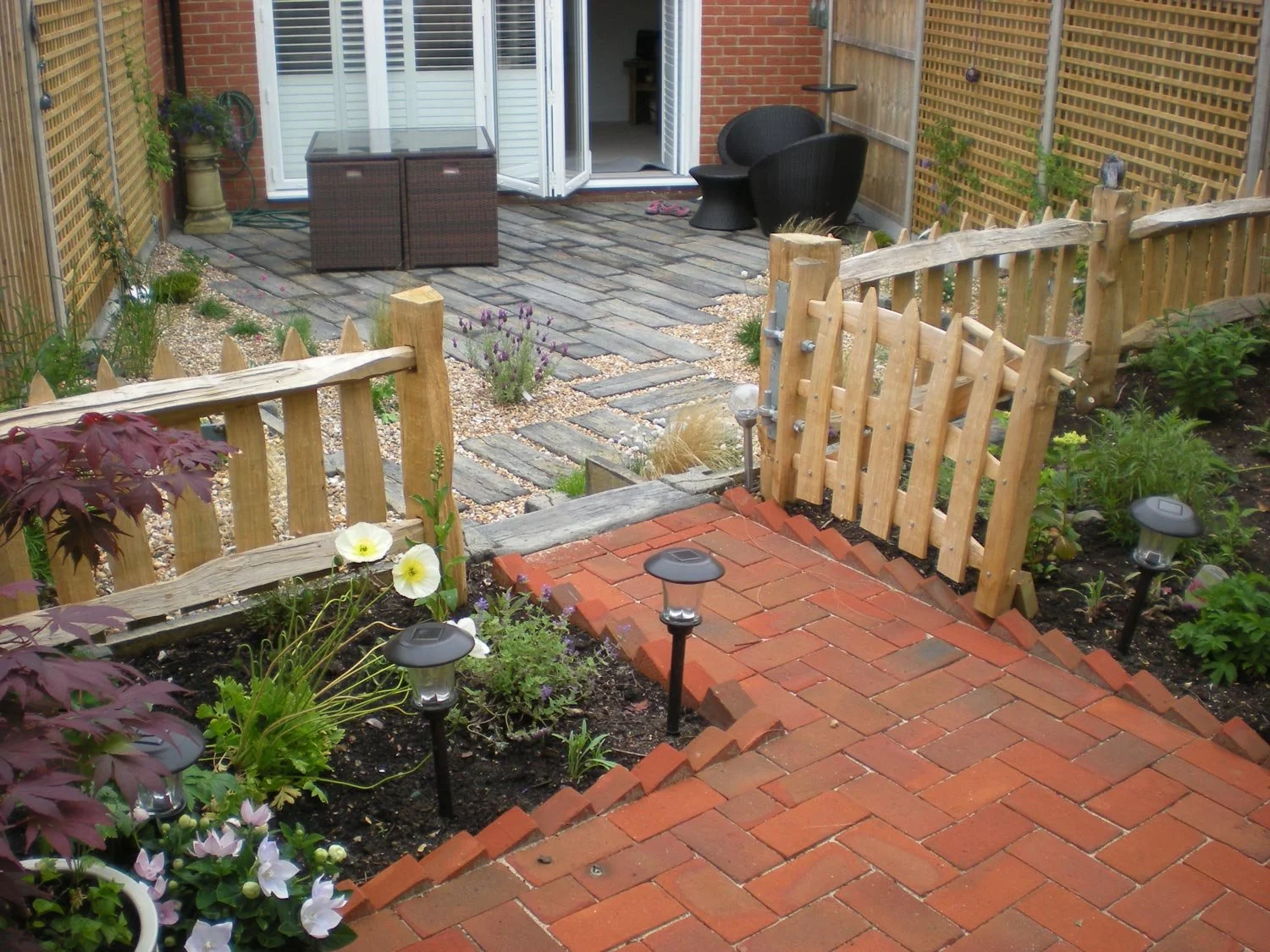 Backyard garden with brick pathway, wooden fences, small garden beds with plants, flower pots, outdoor seating, and a patio with a table and chairs.