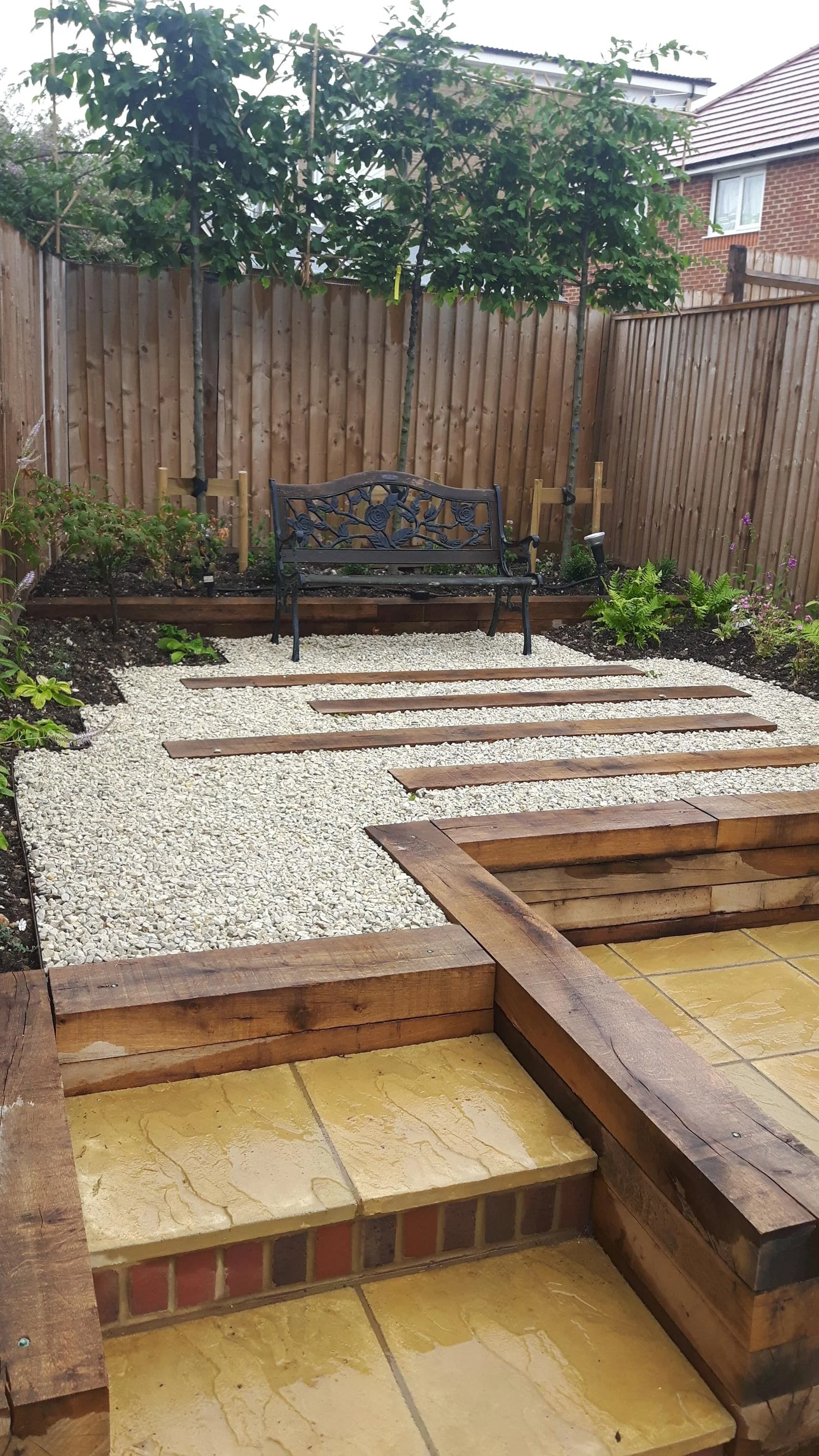 A small backyard garden with a black metal bench, a wooden fence, a planted tree, and flower beds, with stepping stones and gravel paths.