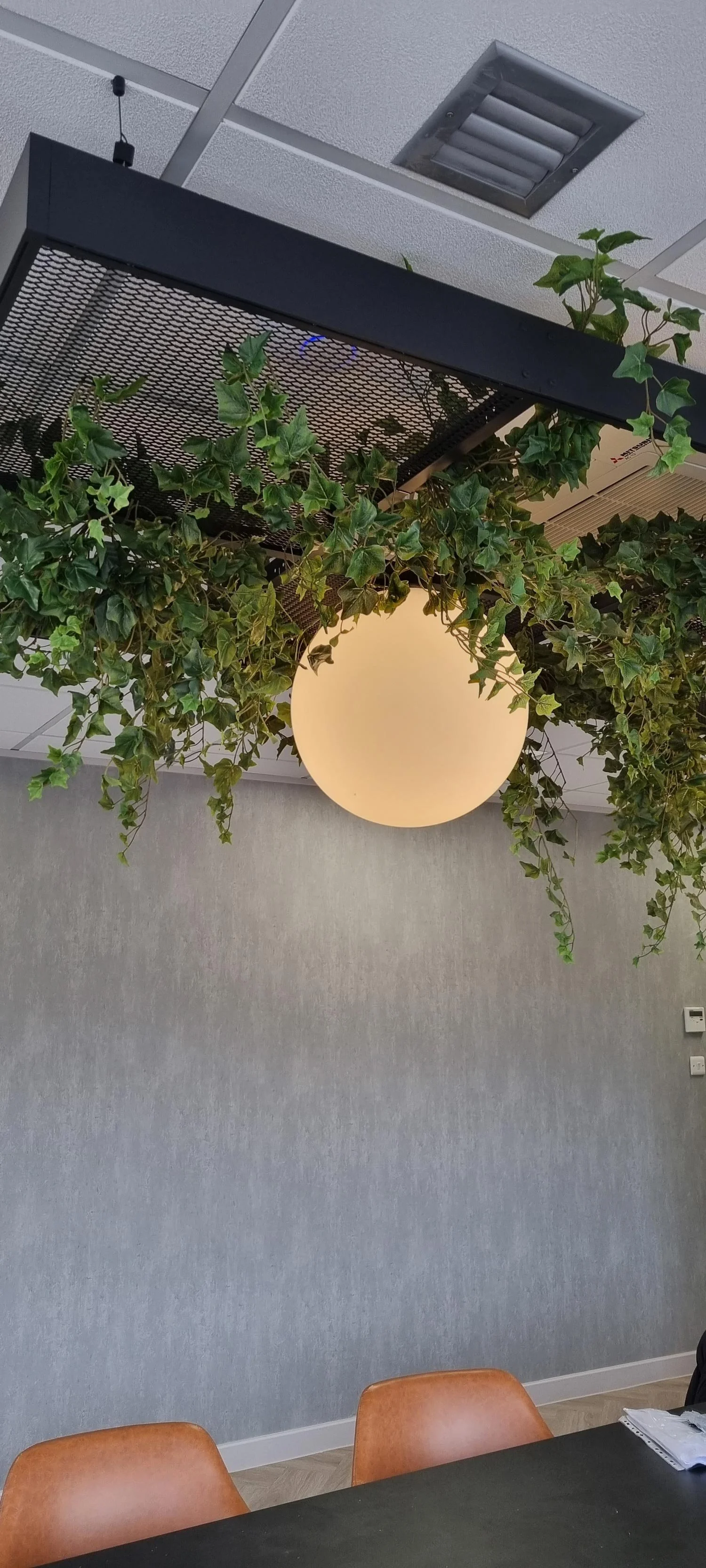 Ceiling with light fixture, greenery, and air vents above an indoor table with chairs in a modern office or meeting room.