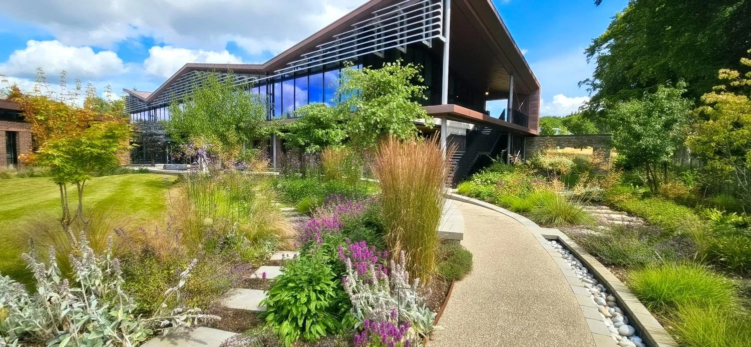 Modern glass building surrounded by lush garden with colorful flowers and green trees under a partly cloudy sky.