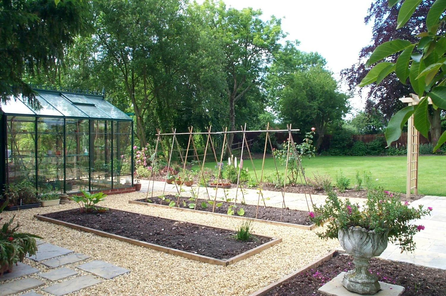 A backyard garden with a greenhouse on the left, raised garden beds with plants, a gravel pathway, lush green grass, trees, a wooden trellis, and flower pots.