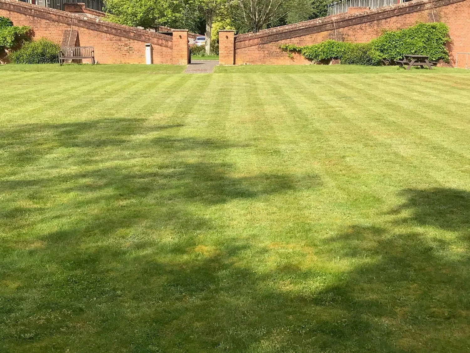A well-maintained grassy lawn with two brick walls and benches, a pathway, and greenery in the background.