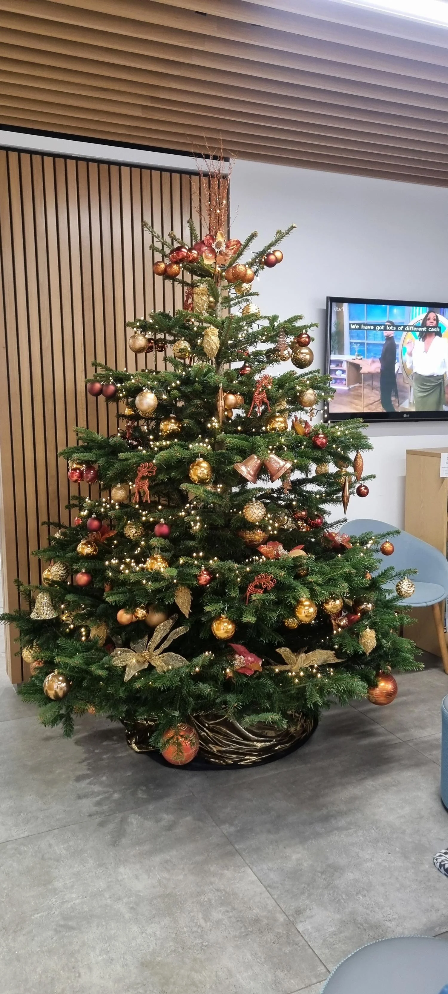 Decorated Christmas tree with gold, red, and bronze ornaments, lights, and gold leaves in a modern indoor setting.