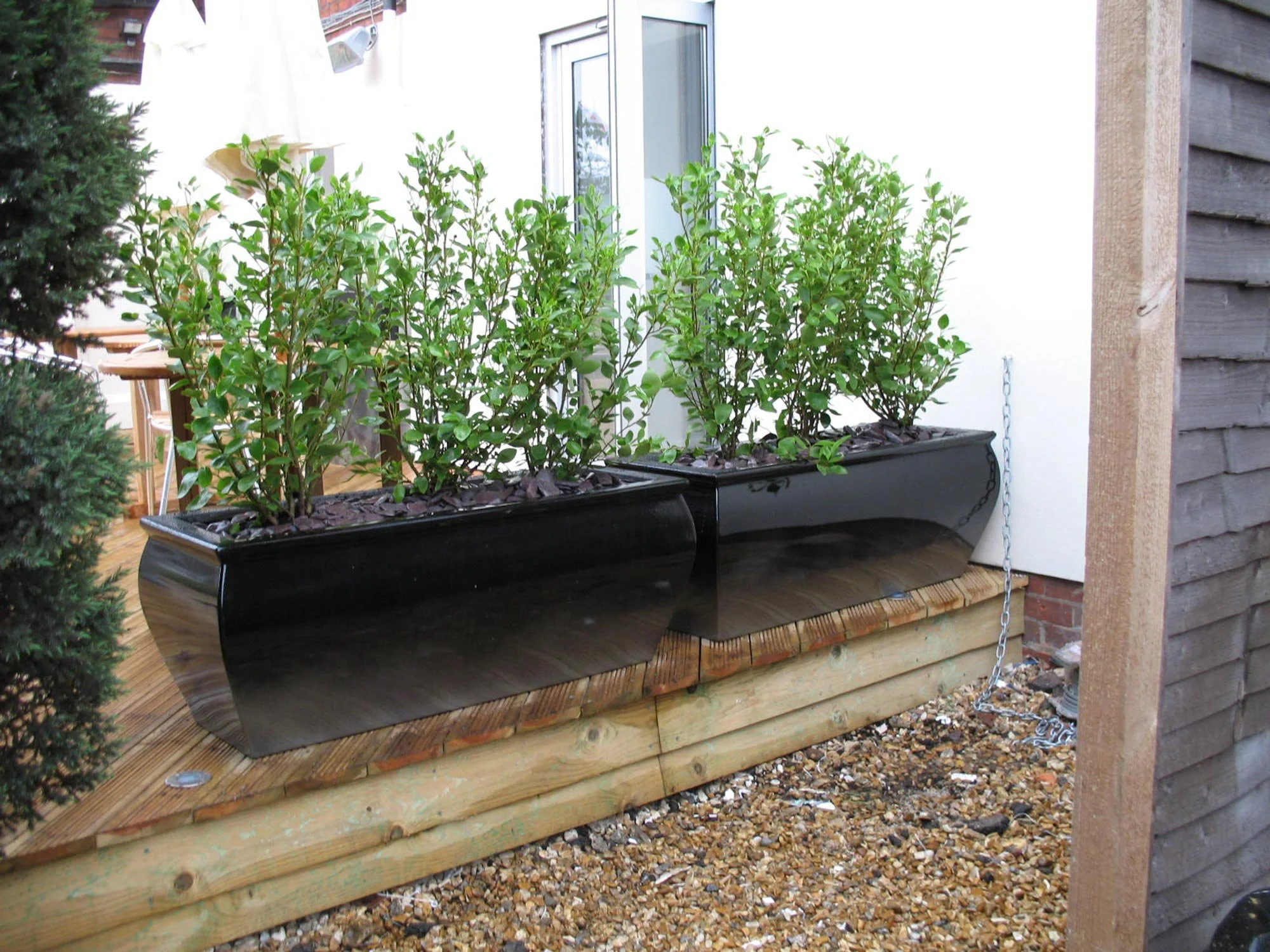 Two black rectangular planters with green leafy plants on a wooden deck next to a white wall, chain attached to the side of the planters, gravel ground underneath, and part of a house with a window visible.