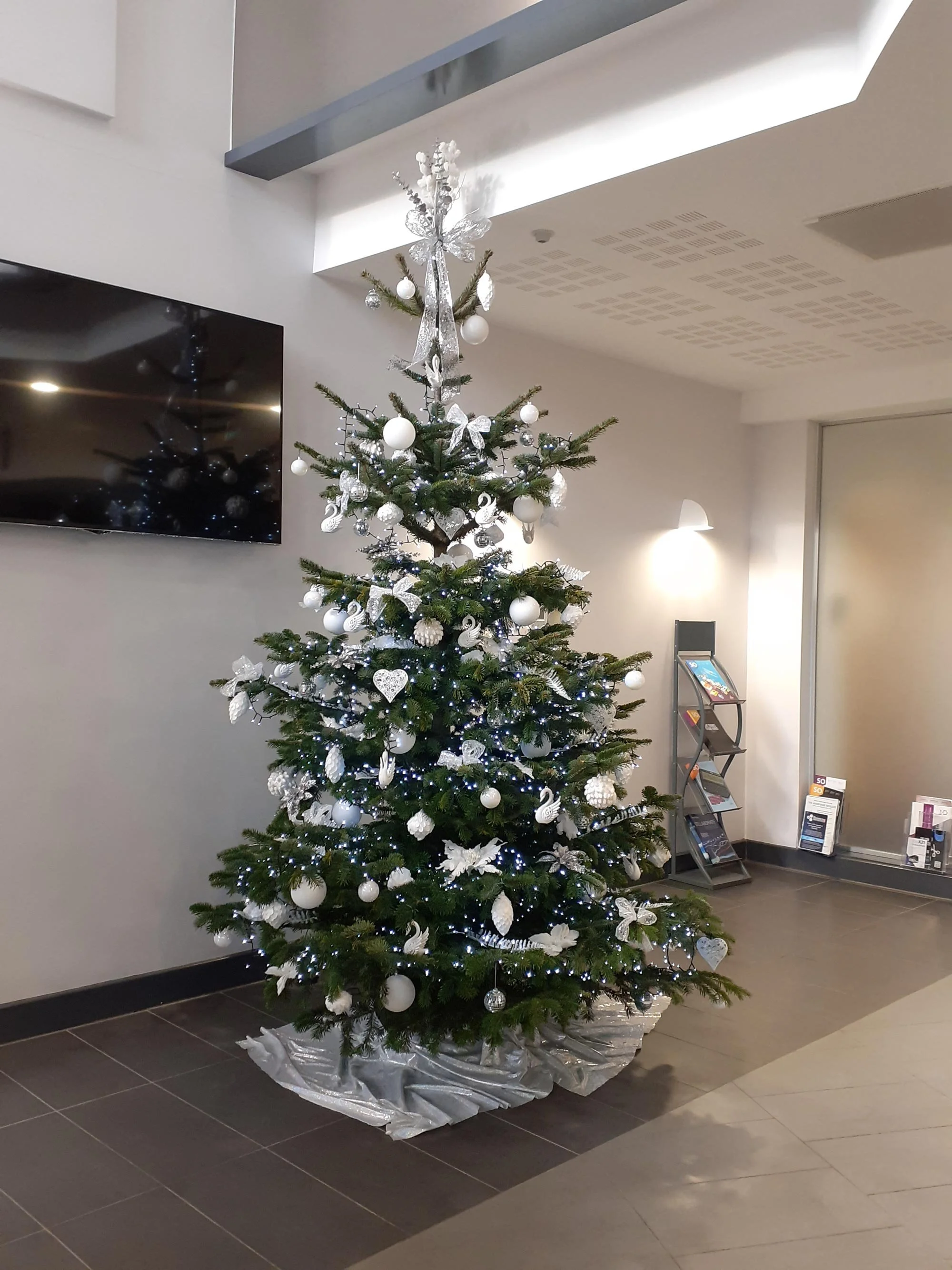A decorated Christmas tree in a modern indoor setting with white and silver ornaments, twinkling lights, and a silver tree skirt.