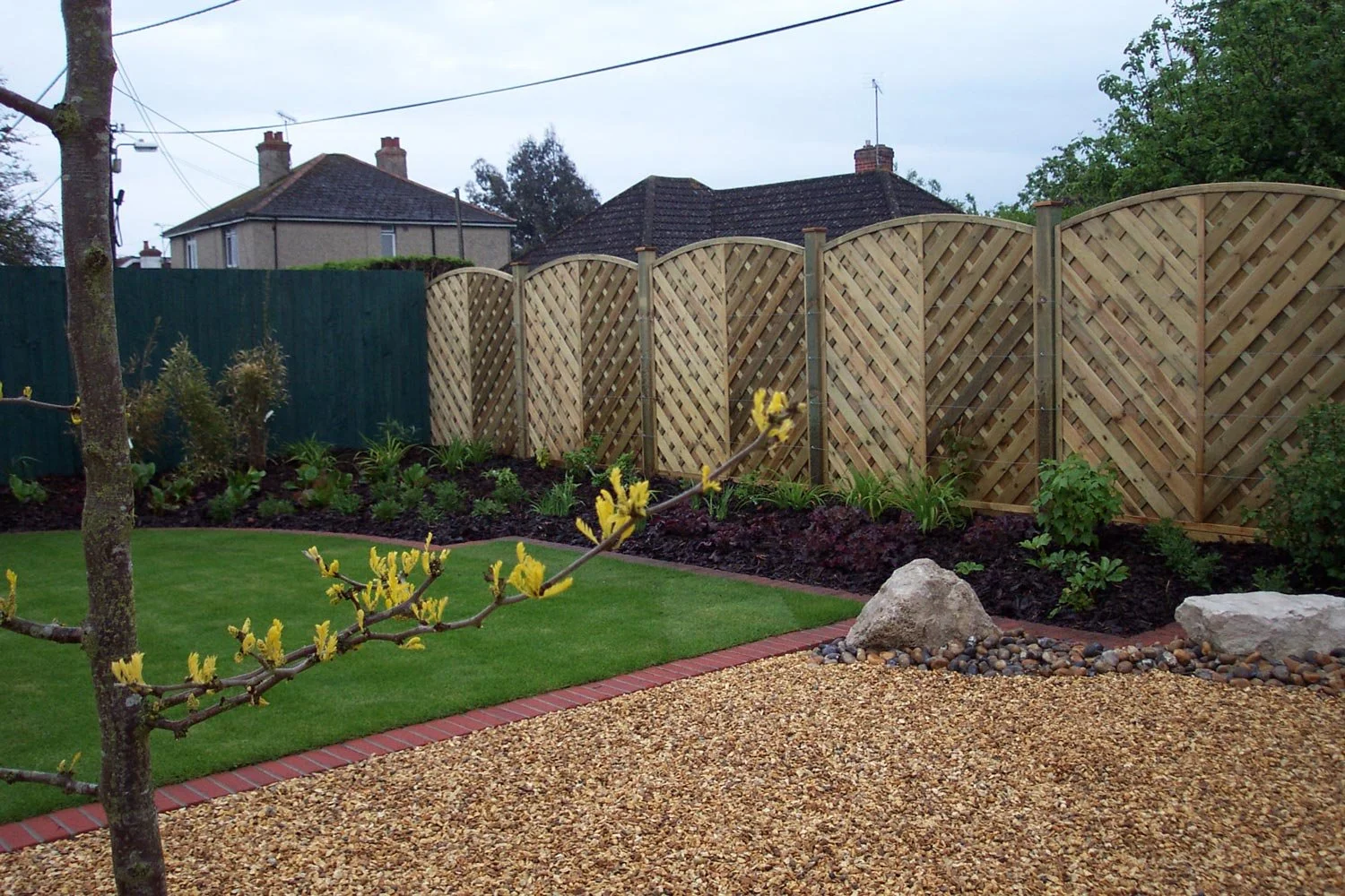 A backyard garden with a neatly trimmed green grass lawn, a gravel area with rocks, a young tree with yellow blossoms, a flower bed with green plants and shrubs, a wooden fence, and houses in the background under a cloudy sky.