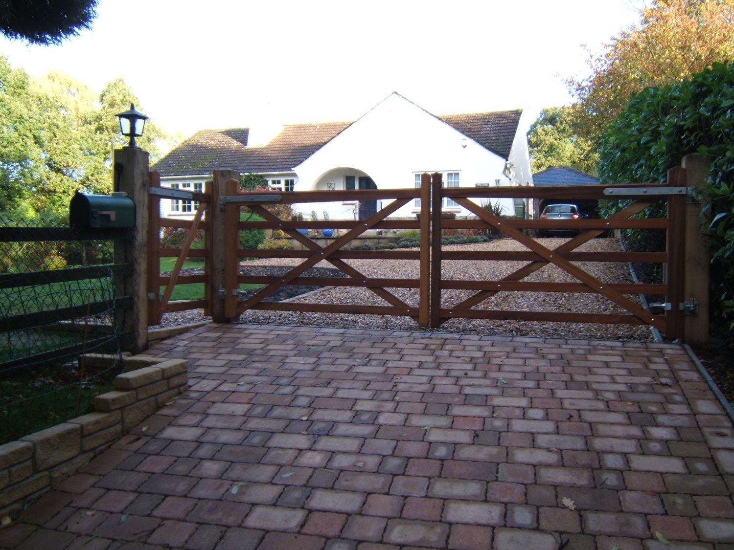 A brick driveway and a closed wooden gate leading to a white house with a gravel driveway and parked car, surrounded by trees and bushes.