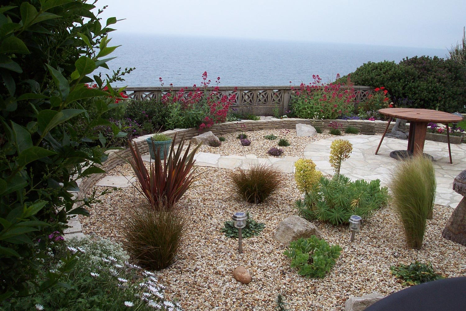 A coastal garden with colorful flowers, desert plants, a stone patio, and a view of the ocean in the background.