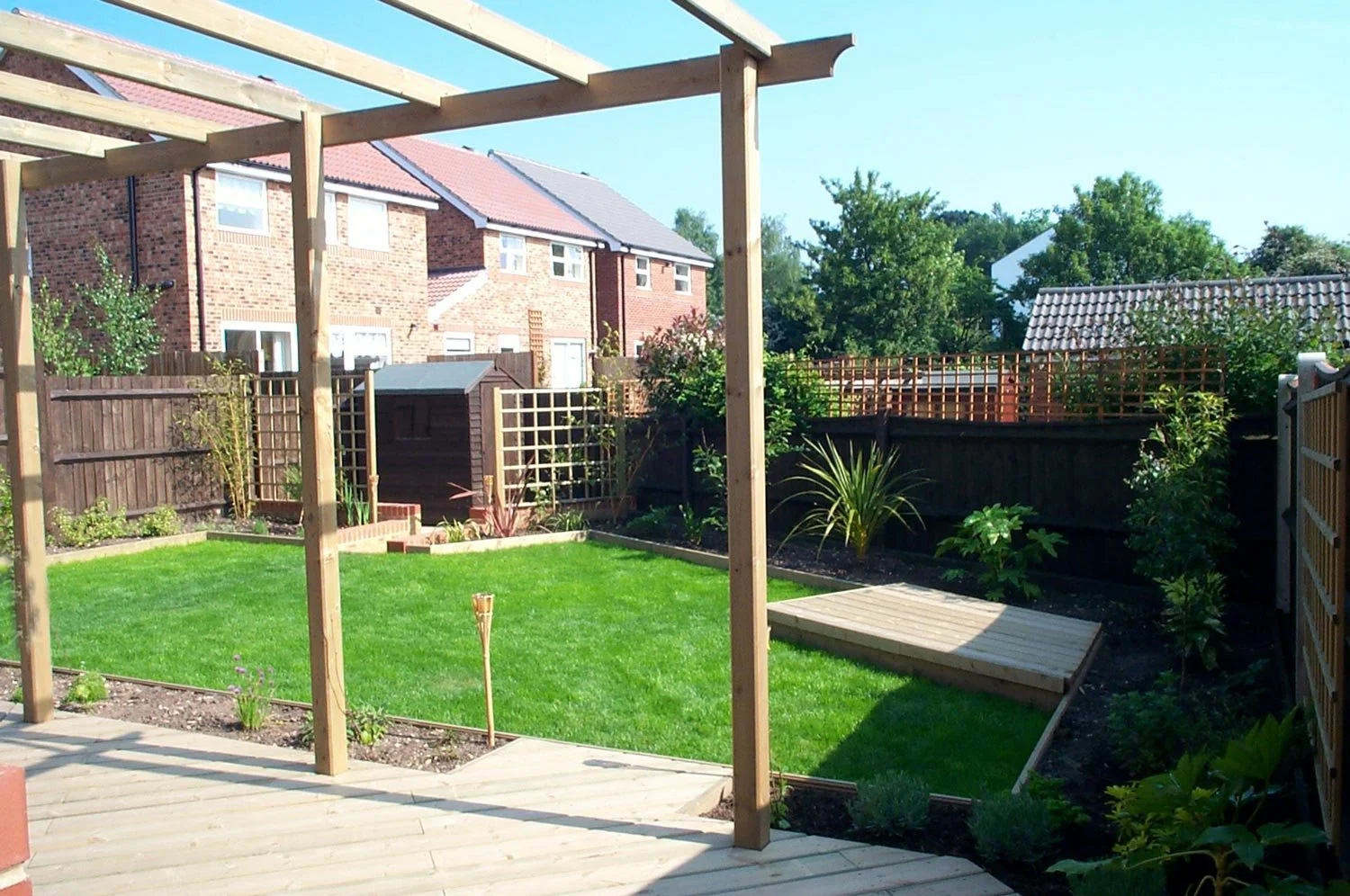 Backyard with freshly laid lawn, small garden bed, and a wooden pergola frame next to a deck.