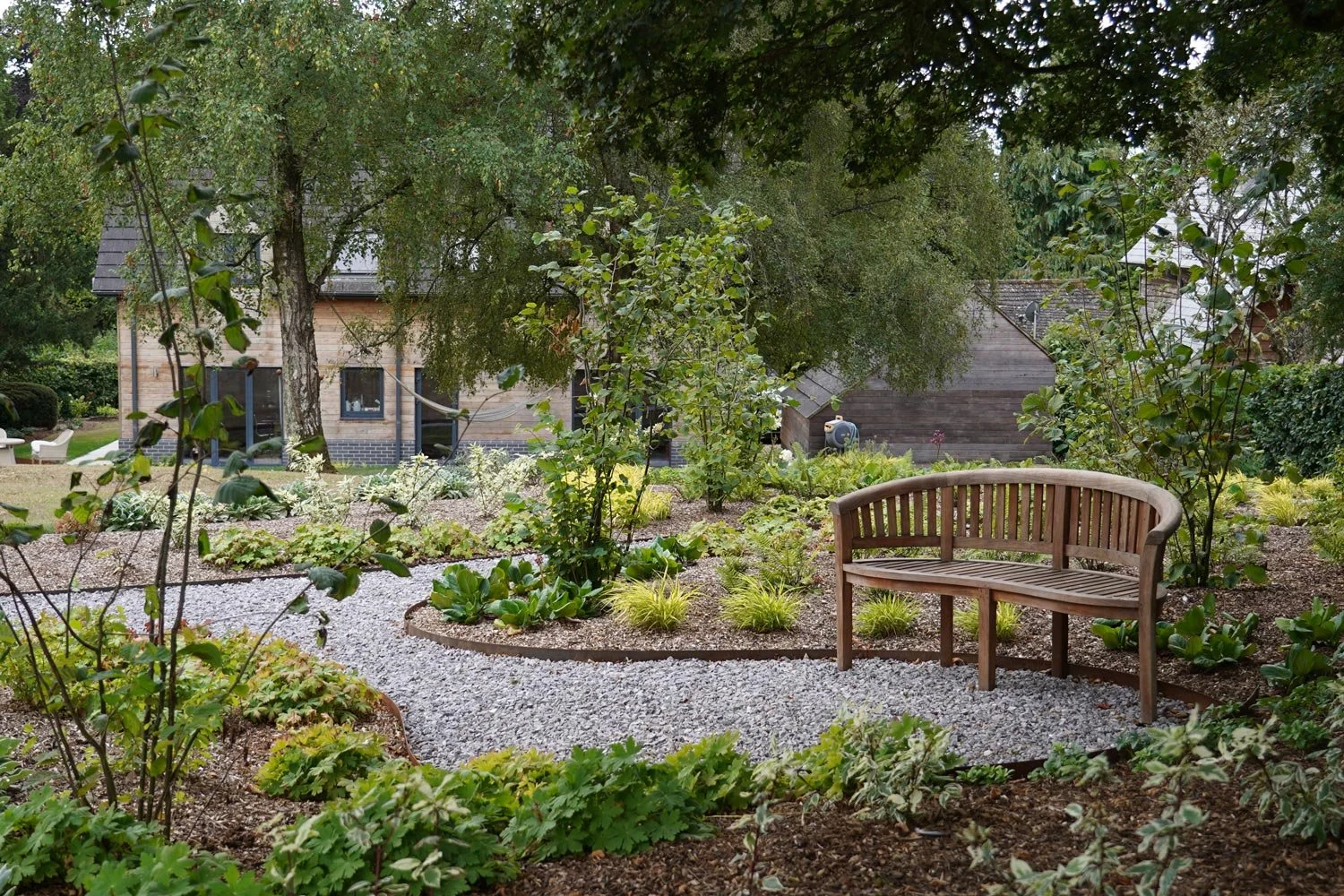 A lush garden with a wooden bench on a gravel pathway, surrounded by various green plants and trees, with a house in the background.