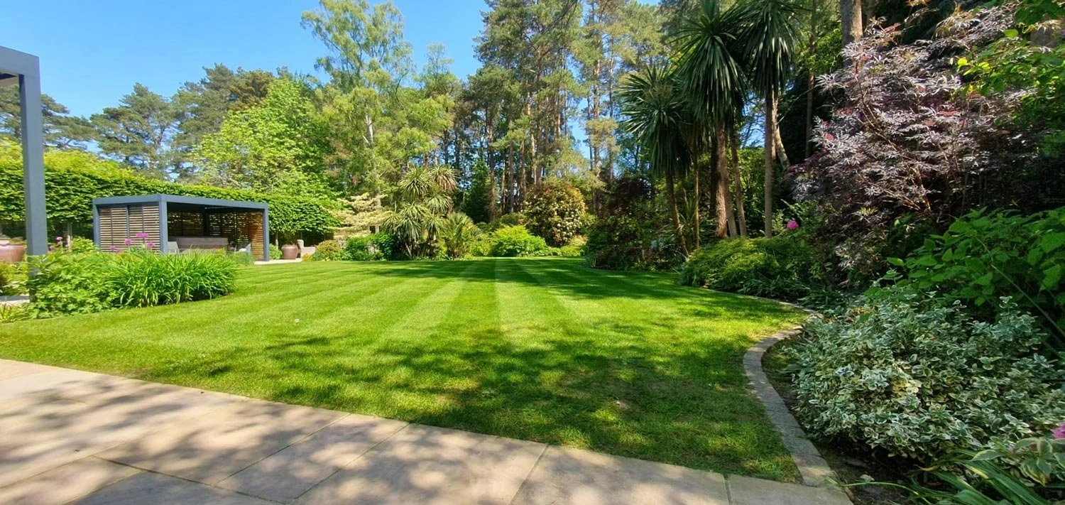 A well-maintained backyard with a lush green lawn, flower beds, trees, shrubs, a paved walkway, and a small outdoor shelter or gazebo.