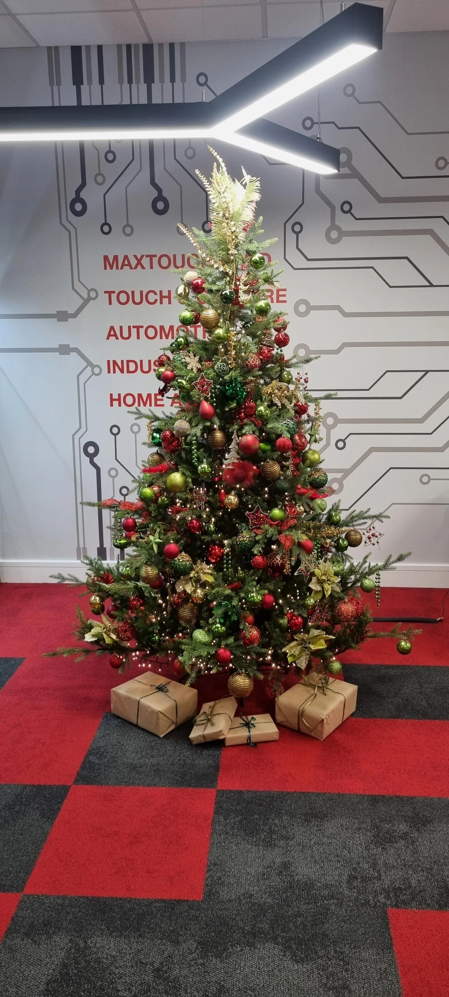 Decorated Christmas tree with red, gold, and green ornaments and wrapped presents underneath, set against a wall with technological-themed graphics.